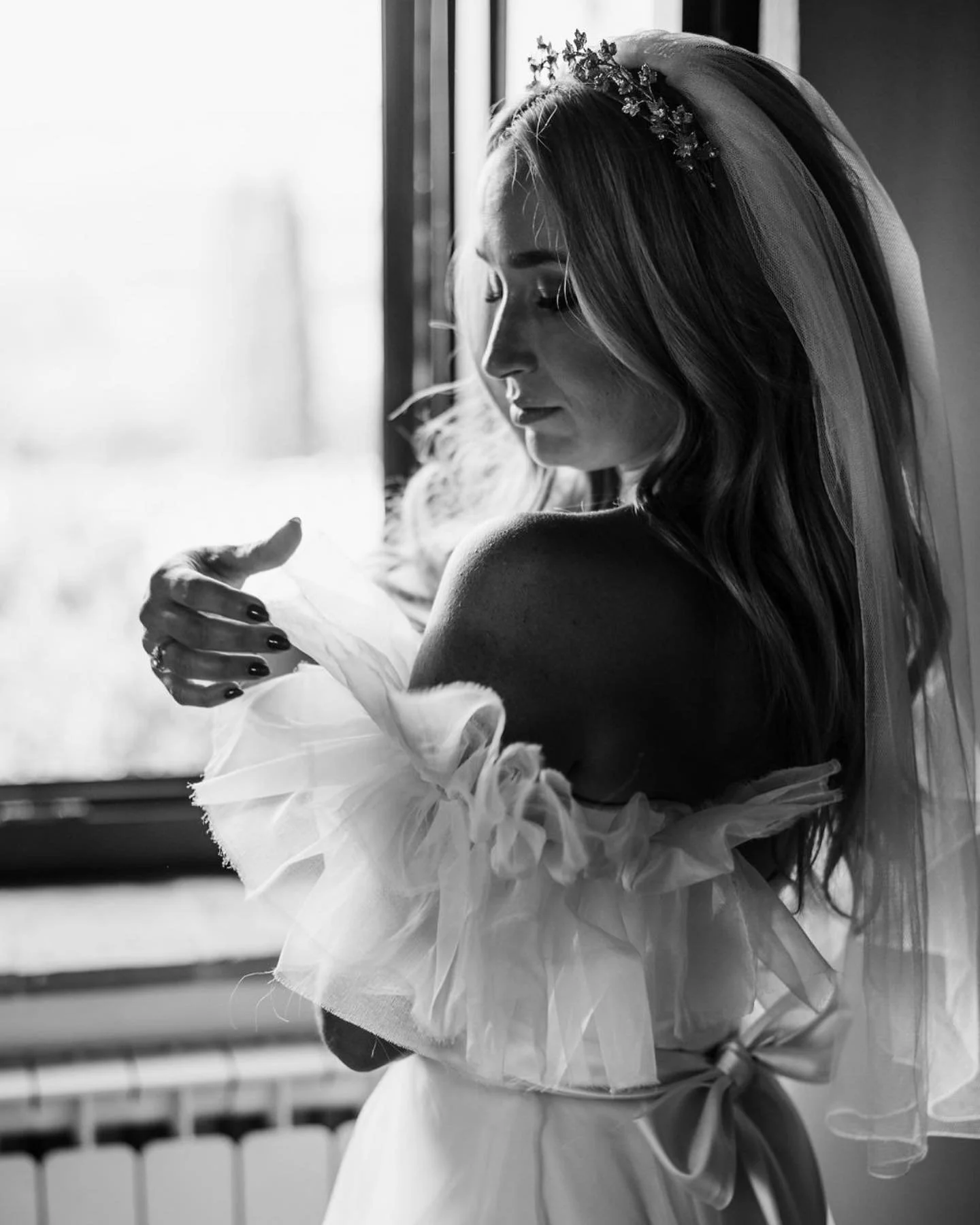 Black and white photograph of a bride with long hair, wearing a veil and floral headpiece. She stands by a window, gently holding her arm close to her chest with her eyes closed.