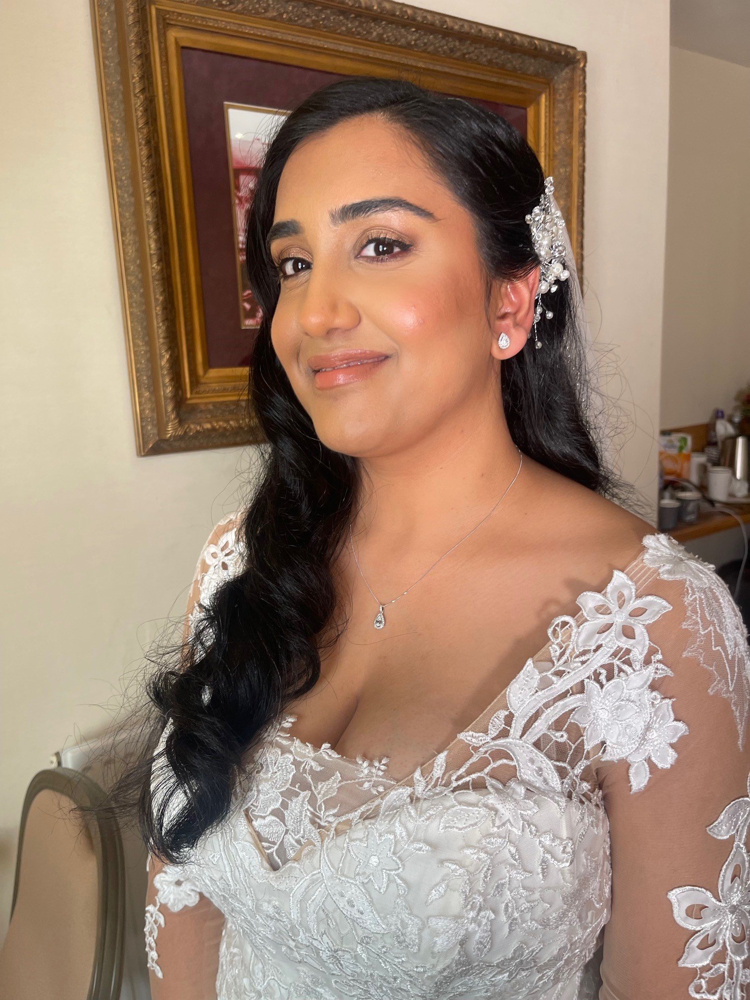 A woman with long black hair and pearl hair accessories wearing a white lace dress with floral embroidery, smiling slightly, standing in a room with a framed picture on the wall behind her.