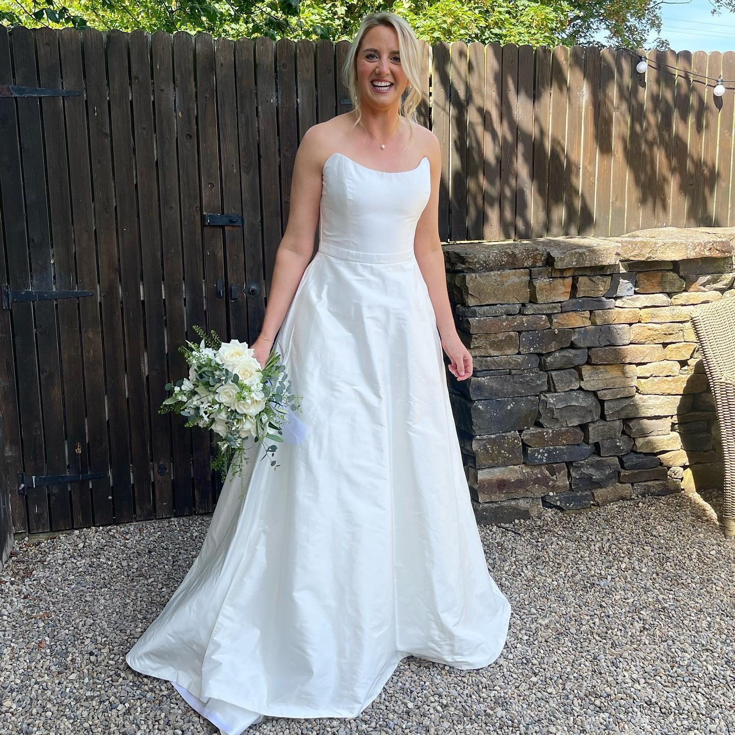A smiling woman in a white wedding dress holding a bouquet of white flowers, standing outdoors on a gravel surface with a wooden fence and greenery in the background.