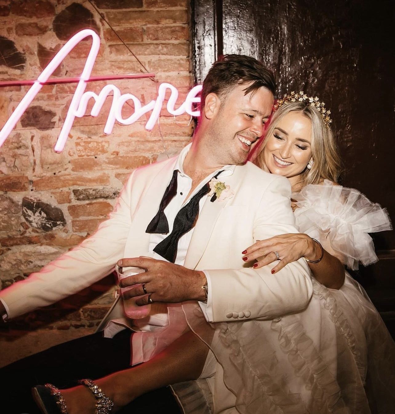 A happy couple at their wedding reception, sitting close together and smiling, with a brick wall and a neon pink 'love' sign in the background.