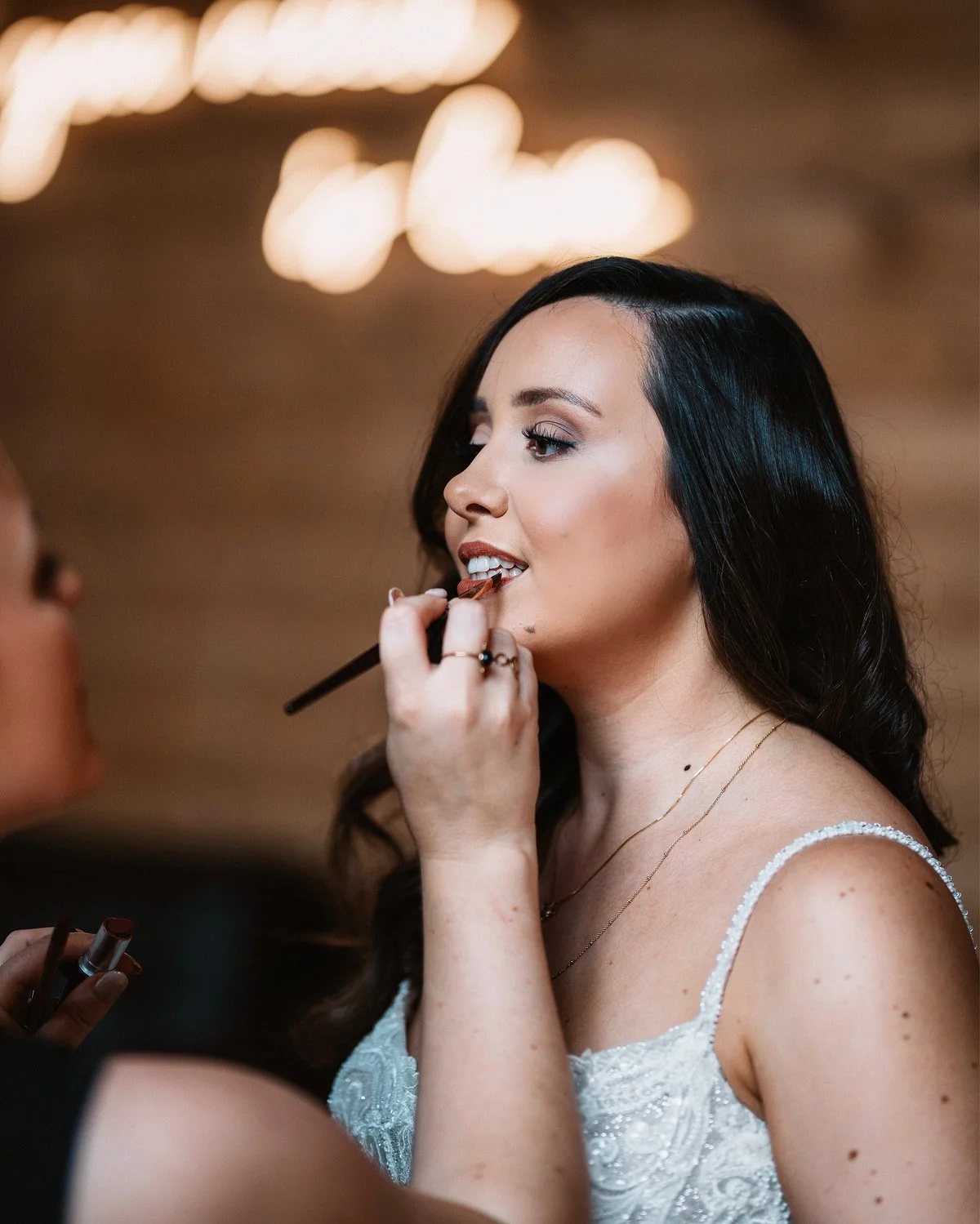 A woman with dark hair and light skin gets her makeup done, with a makeup artist applying lipstick. She wears a white dress with thin straps and a delicate necklace.