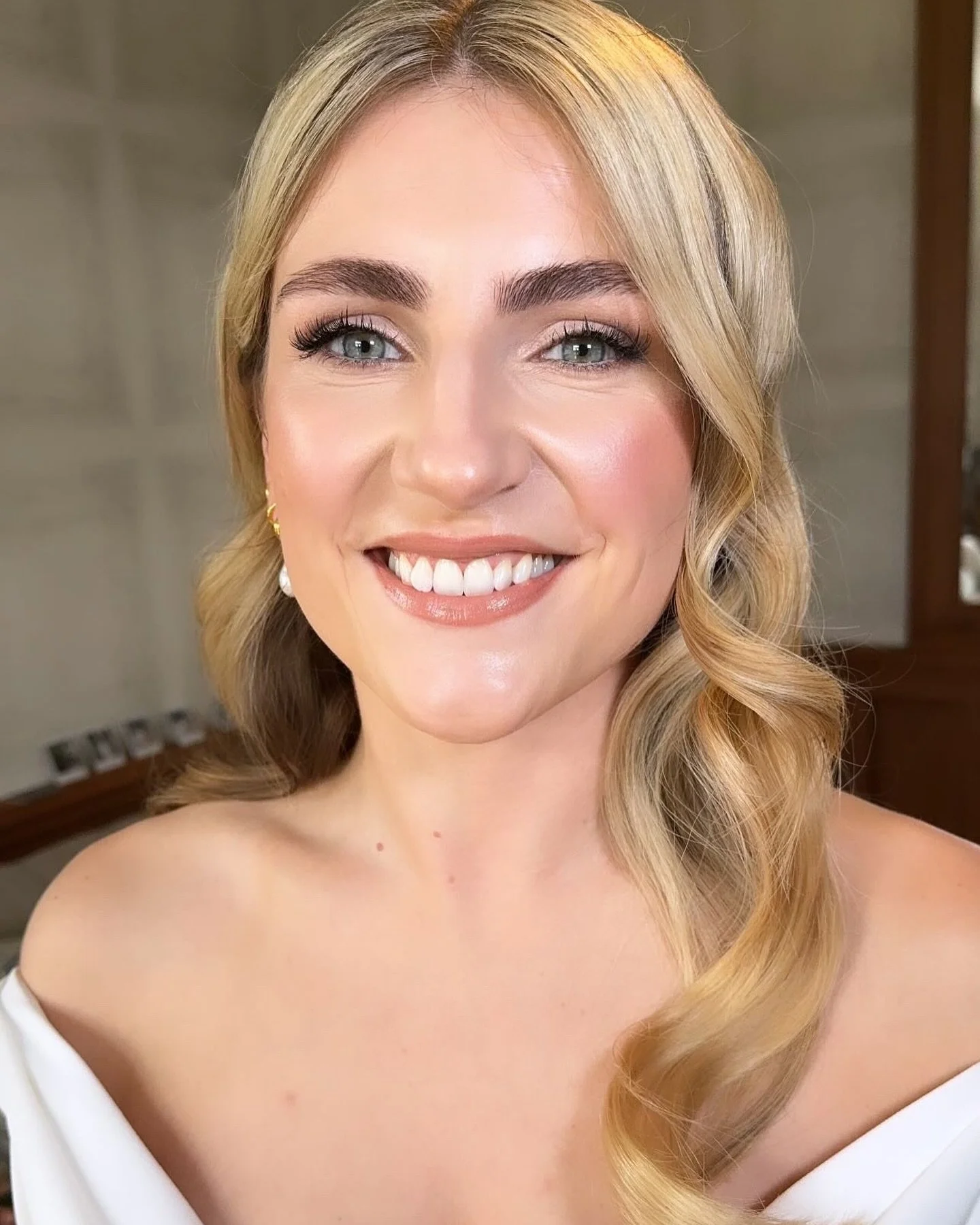 A smiling woman with fair skin, blonde wavy hair, blue eyes, and makeup, wearing a white off-shoulder top, indoors with blurred background.