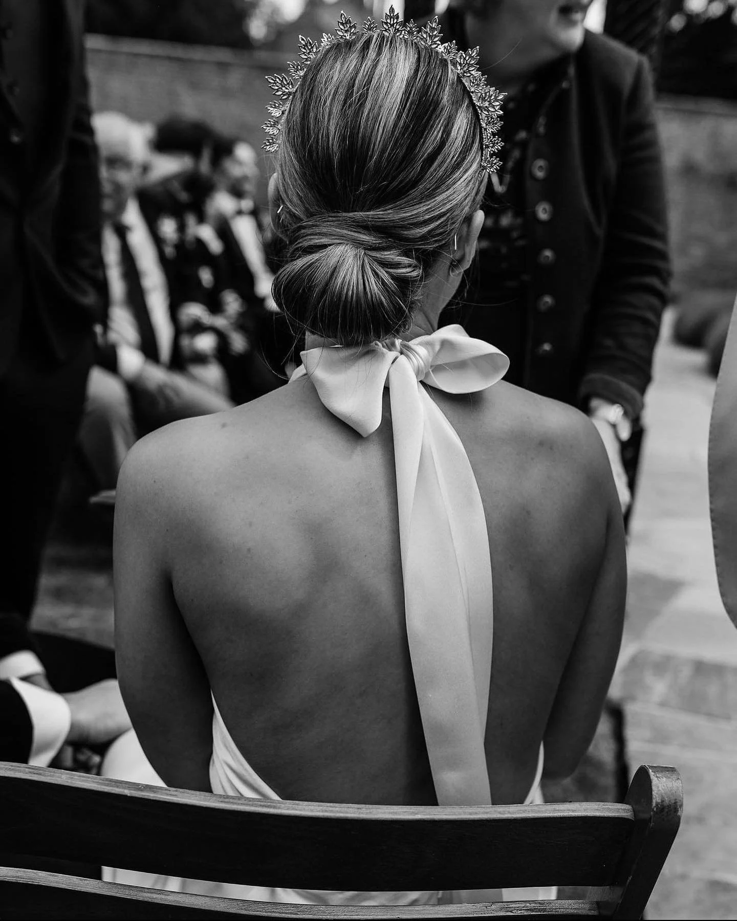 A woman at a wedding or formal event with her back to the camera, wearing a decorative crown and a scarf tied around her neck, sitting on a wooden bench.