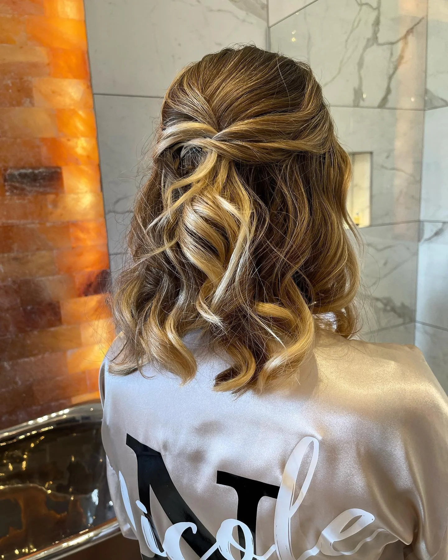 Back view of a woman with shoulder-length, curly blonde hair styled with loose curls and a twisted section on top. She is wearing a white satin or silk garment with text on the back, possibly at a salon or similar setting with a marble wall and a woo