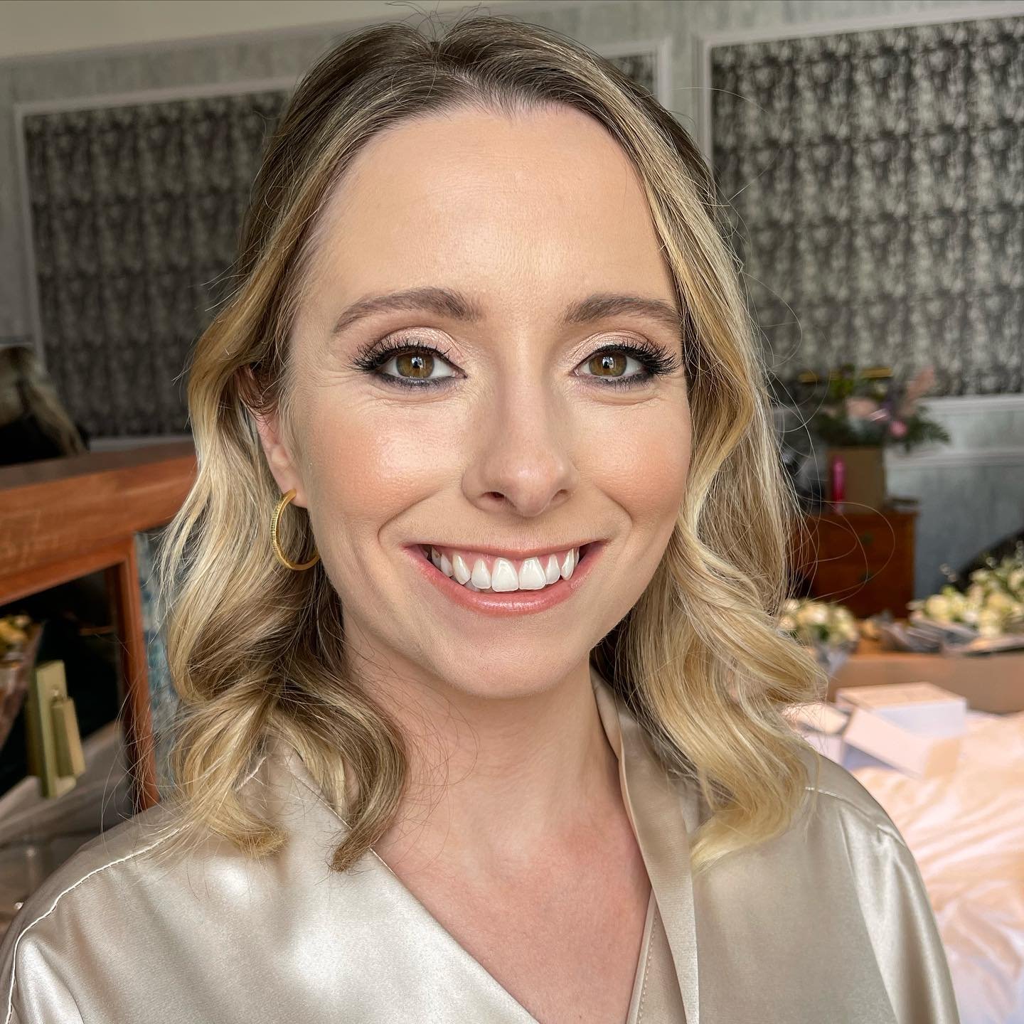 A woman with light brown hair styled in loose curls, wearing hoop earrings and a beige satin robe, smiling in a room with floral curtains and a table with flowers and boxes in the background.
