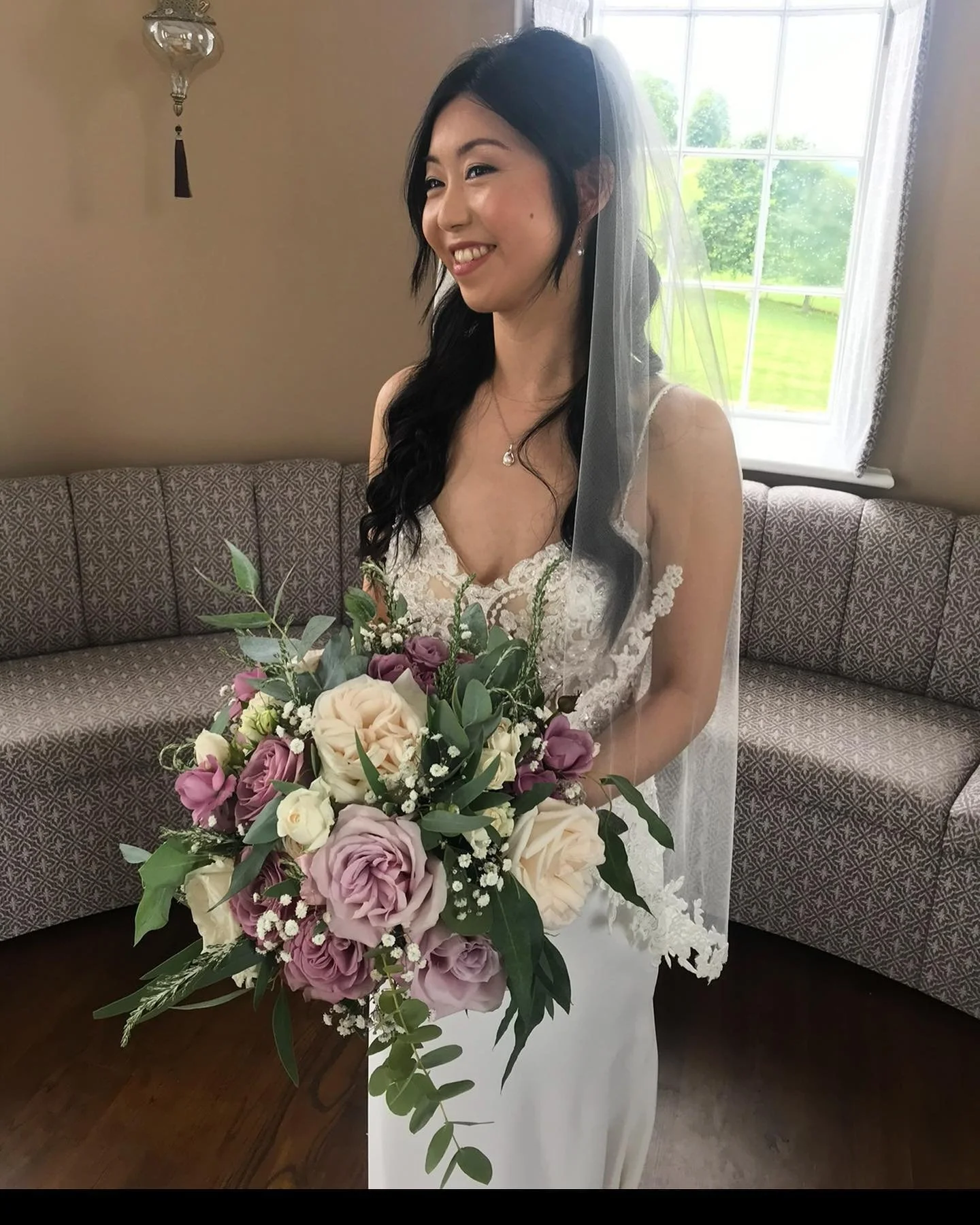 A bride in a white lace wedding dress, holding a large bouquet of pink, purple, and cream roses with greenery, smiling inside a room with a window and patterned sofa in the background.