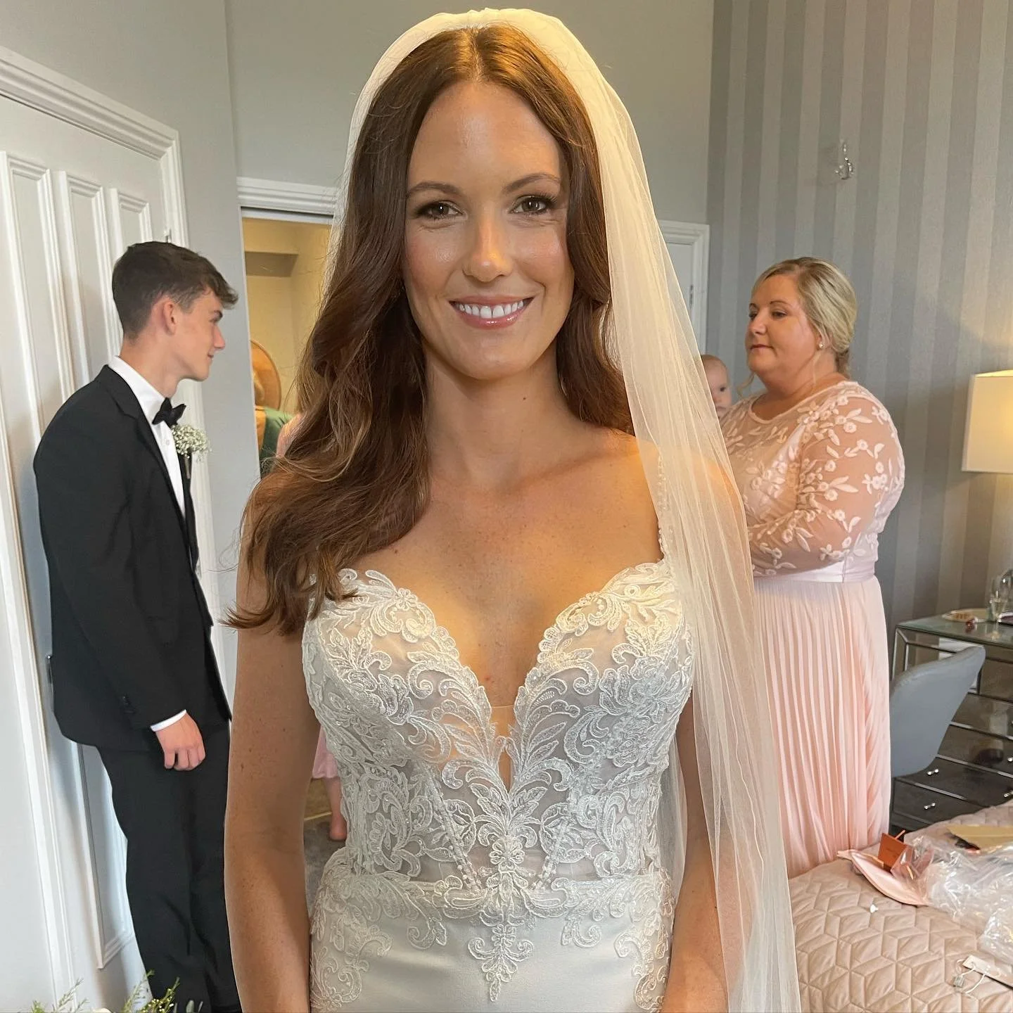 Bride in a wedding dress with lace details, smiling, with a veil, in a room with a man in a tuxedo and a woman in a pink dress in the background.