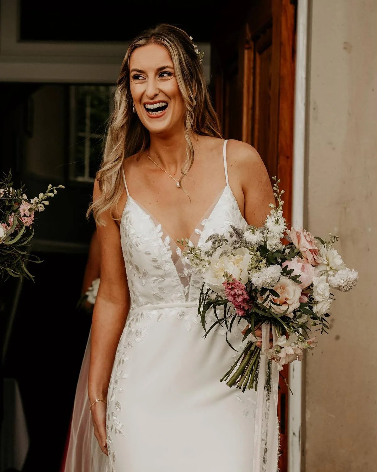 A woman in a white wedding dress holding a bouquet of pastel pink and white flowers, smiling and looking to the side in a rustic setting.