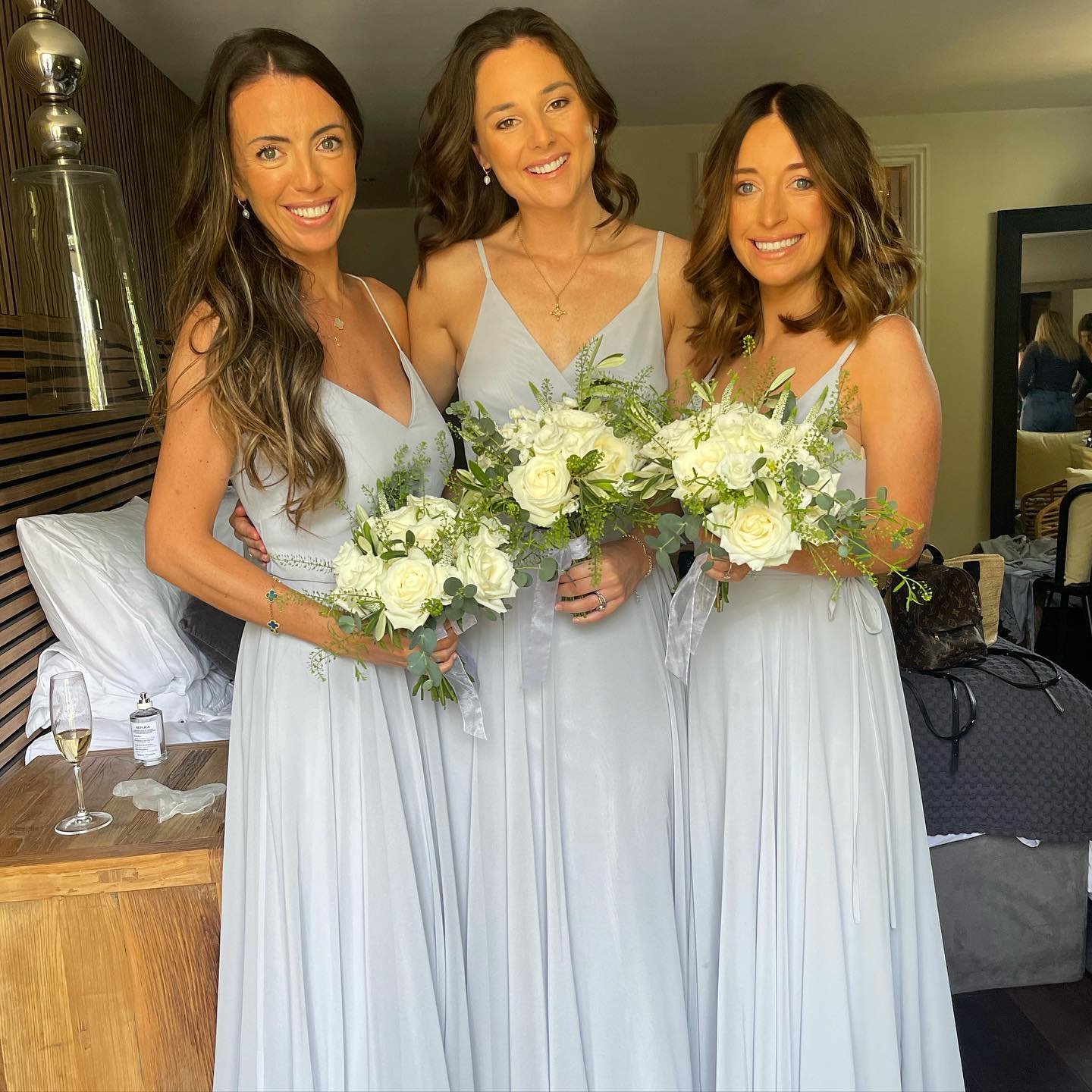 Three women in light blue dresses holding bouquets of white roses and greenery, smiling at the camera indoors.