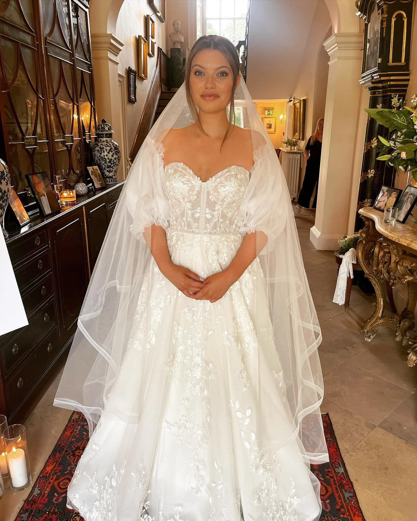 A bride in a white wedding dress with lace details, standing indoors with her hands clasped in front. She is wearing a sheer veil and has her hair styled in an updo. The background shows a staircase, framed photographs, and decorative vases in a well