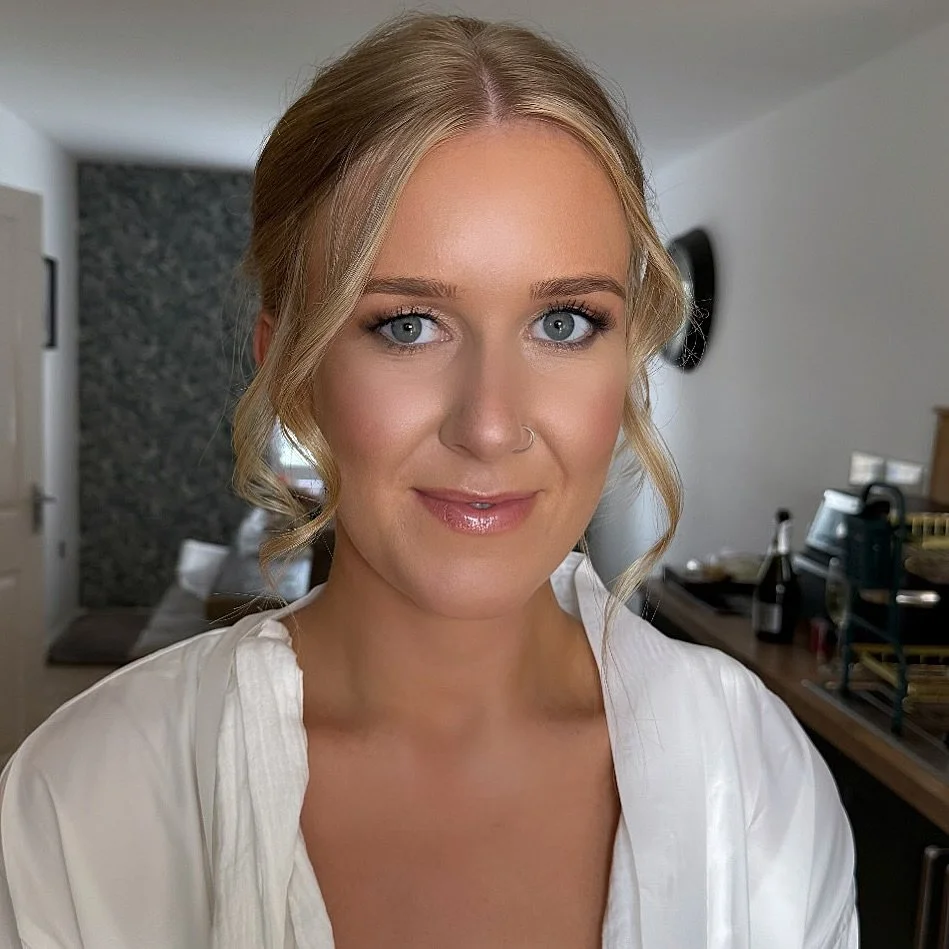 A woman with blonde hair styled in loose waves, blue eyes, and wearing a white top. She has a small nose piercing and light makeup with glossy lips. The background shows a modern living room with a gray accent wall, a door, and a table with various i