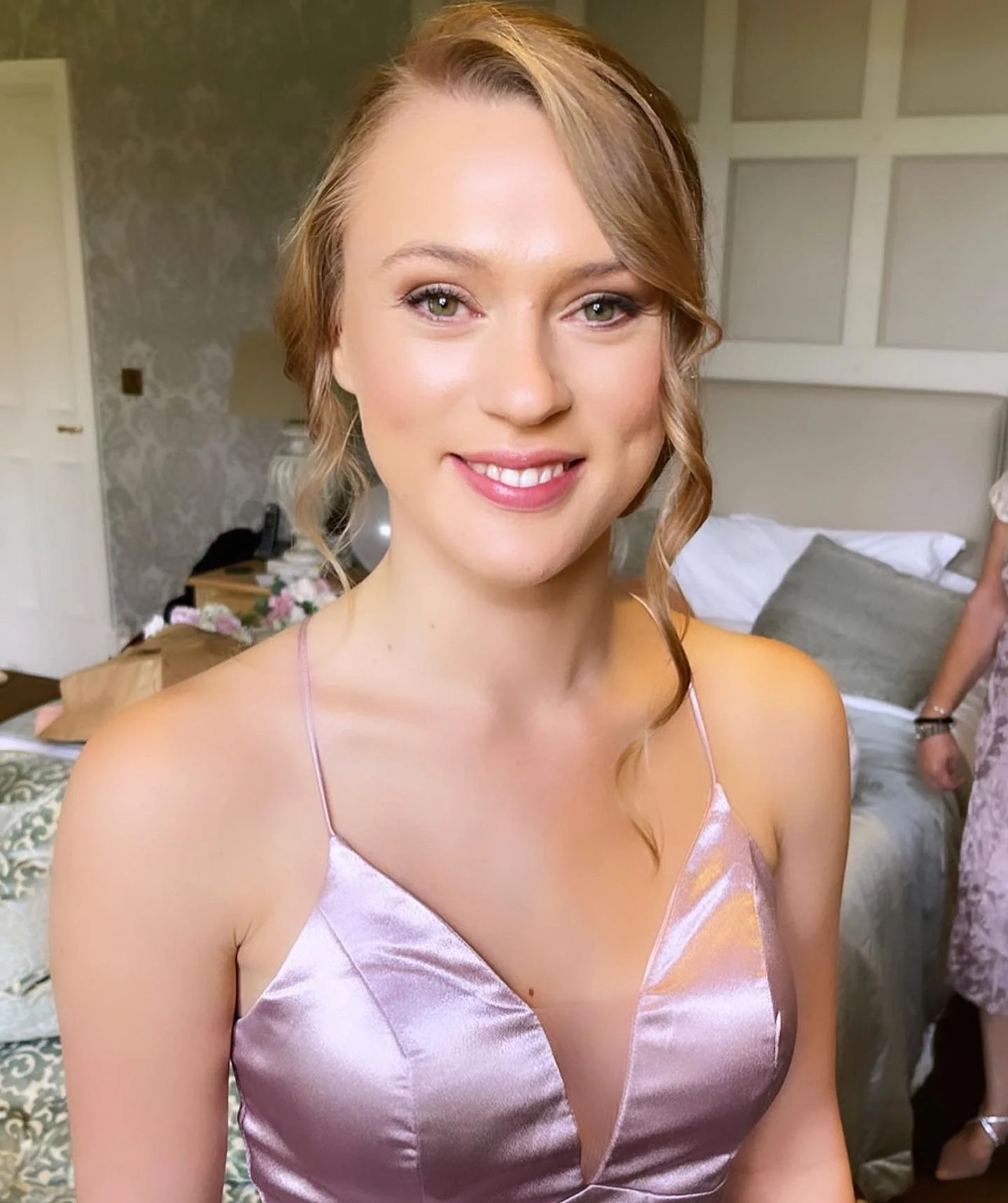 A woman with wavy blonde hair and light green eyes wearing a pink satin spaghetti strap dress, smiling at the camera indoors with a bed and floral decorations in the background.