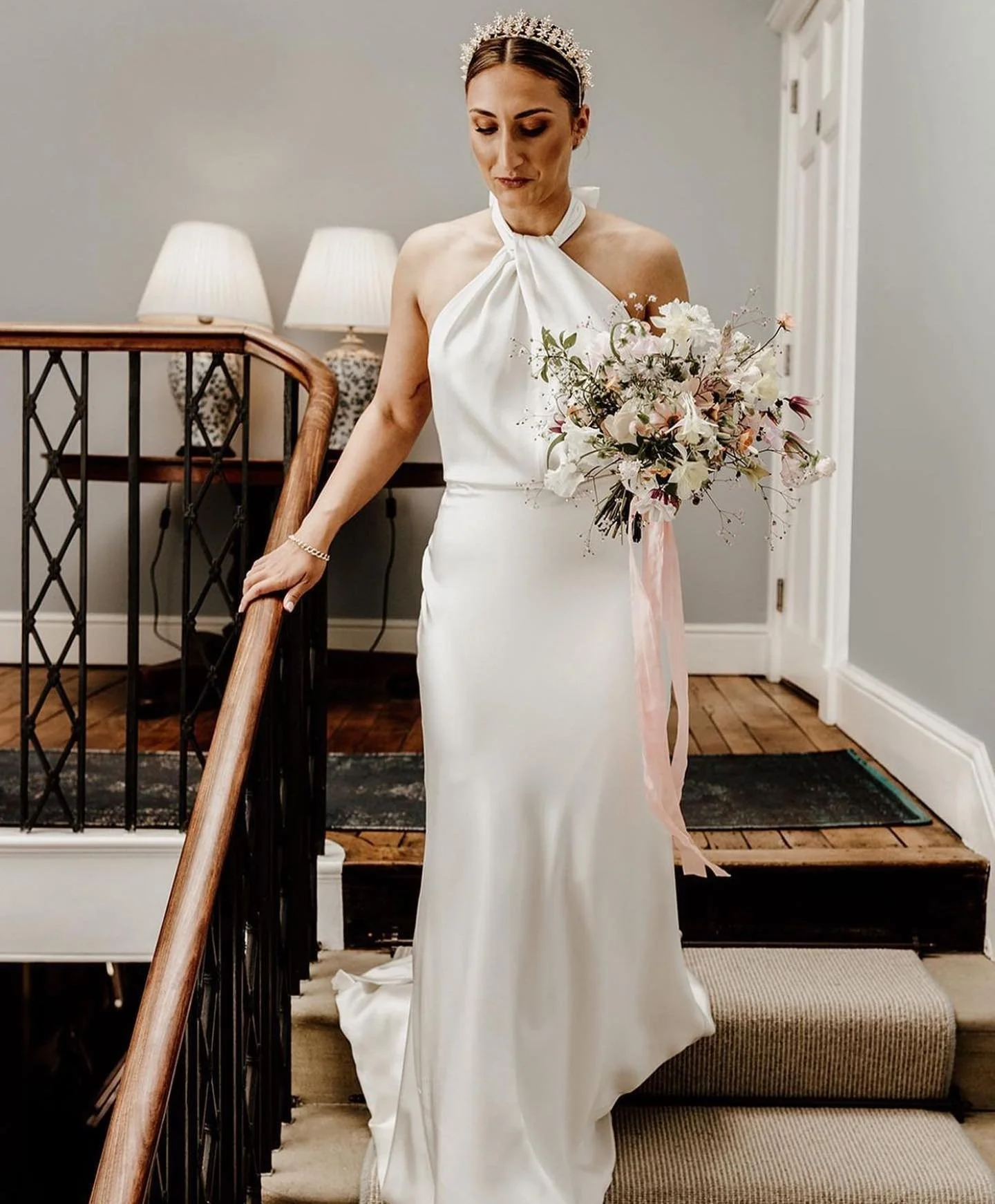 A bride in a white dress holding a bouquet of flowers, standing on a staircase inside a house with a gray wall and two lamps in the background.