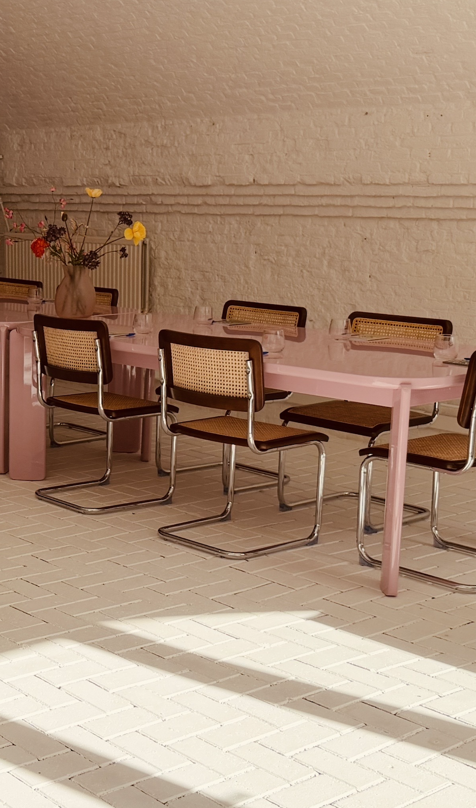 A pink dining table with brown and cane-back chairs in a room with a white brick wall and tiled floor. A vase with colorful flowers decorates the table.
