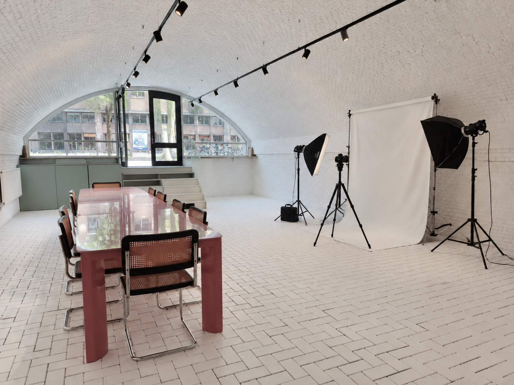 Empty photo studio with white brick walls, a large window, a pink conference table with six chairs, professional lighting equipment, a white backdrop, and some decorative objects on the window sill.