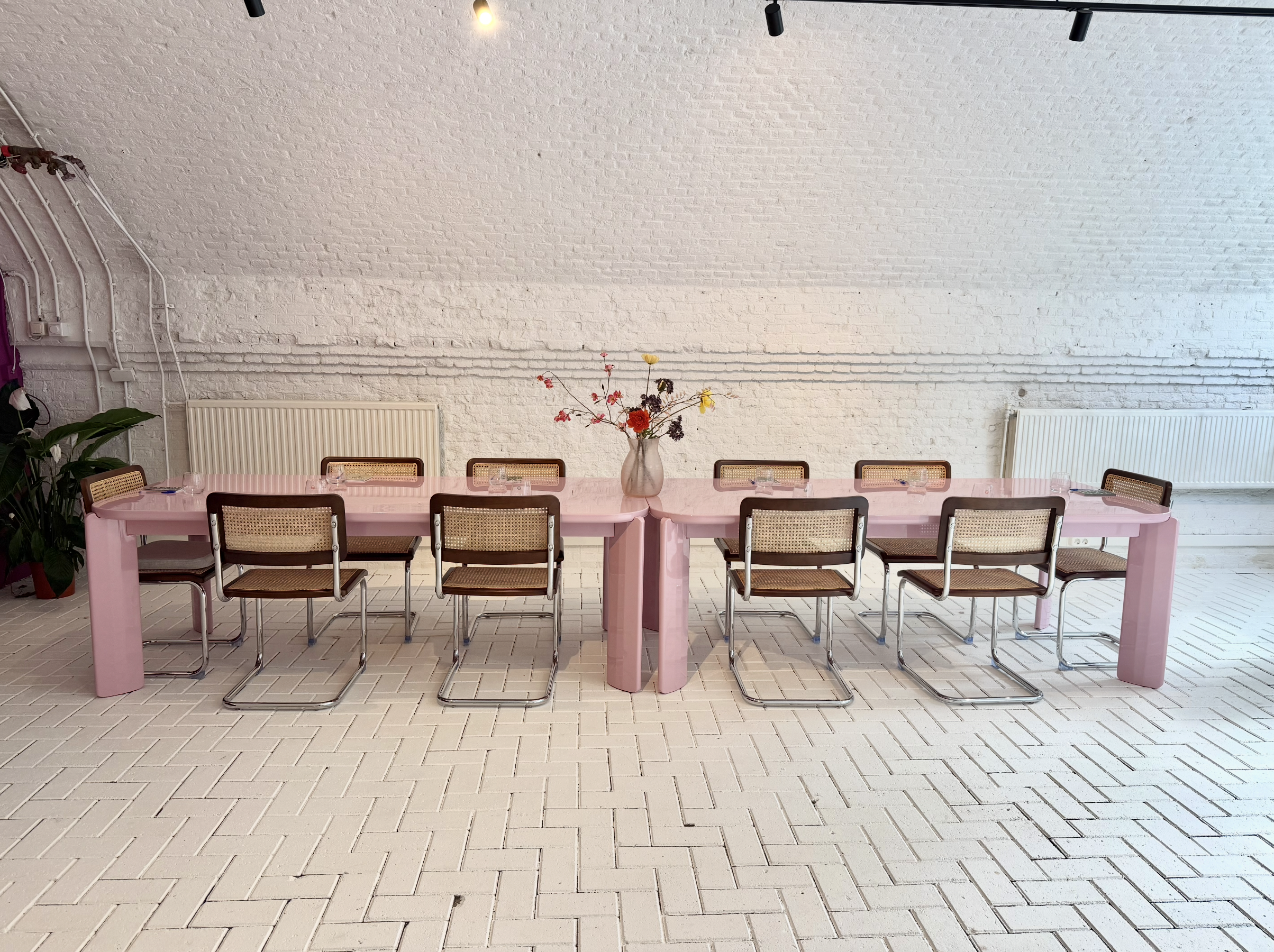 A pink rectangular dining table with a glass vase of colorful flowers in the center. Eight chairs with rattan backs and brown seats are arranged around the table. The background features a white brick wall with a radiator, a large plant on the left, and a plain floor.