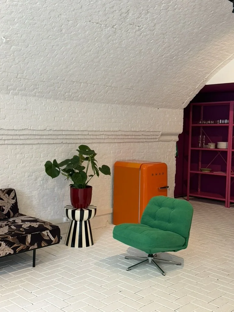 Interior with white painted brick walls and floor, featuring a green lounge chair, a patterned sofa, a potted plant on a striped stand, an orange vintage refrigerator, and a pink shelving unit.