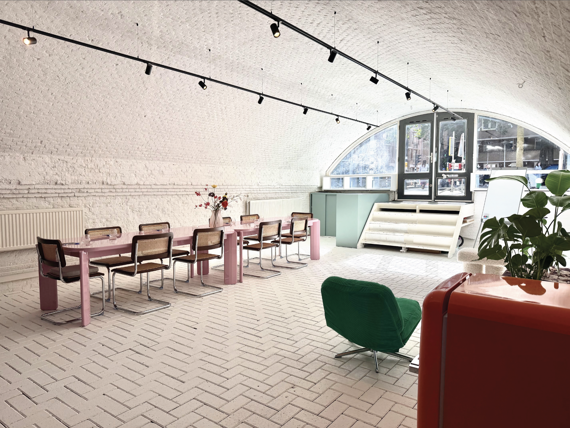 Interior view of a bright, modern room with a white brick wall, pink dining table, several chairs, a green armchair, a red table, and large windows with city street view.