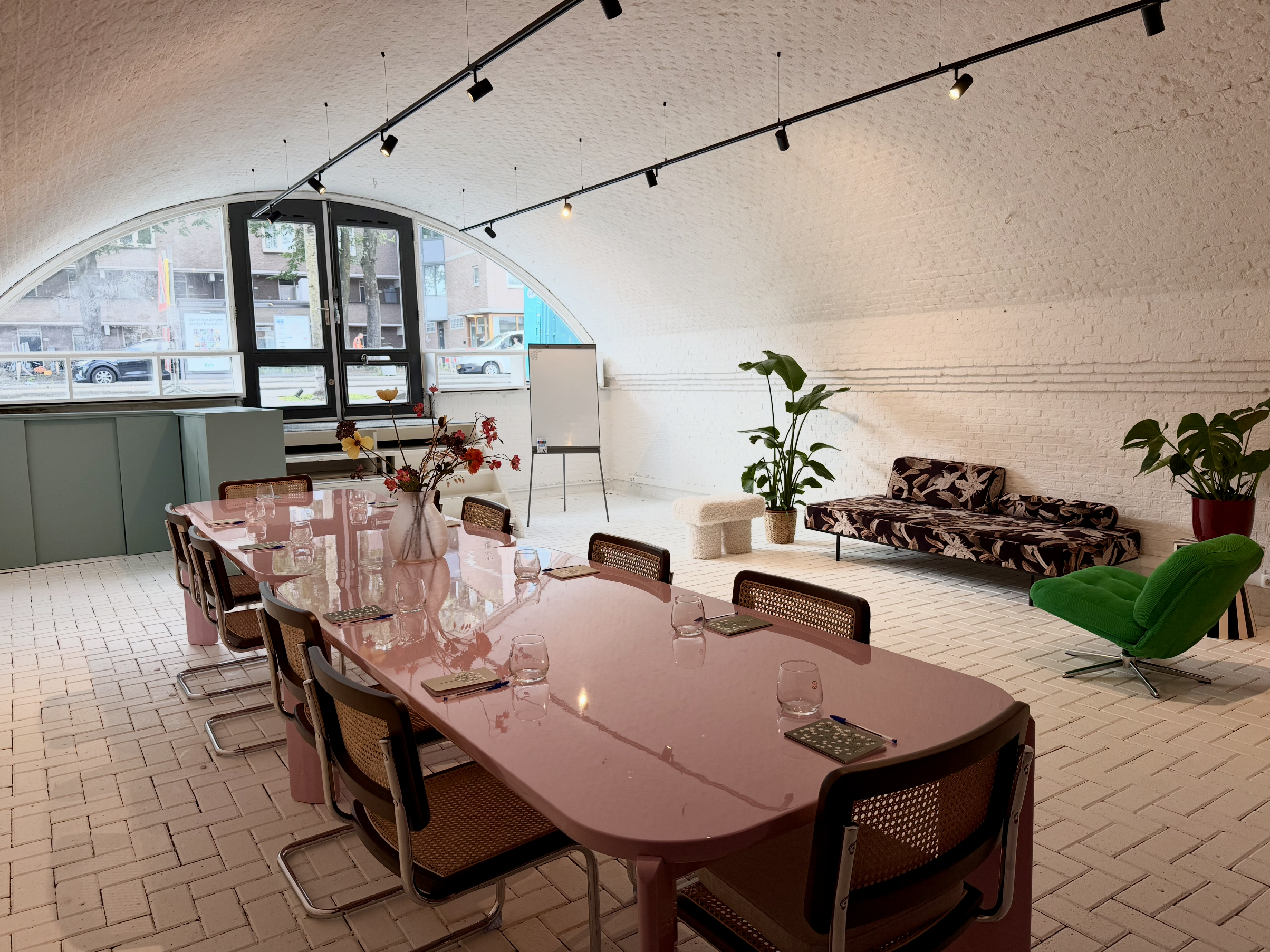 Interior of a modern meeting room with a pink conference table, brown chairs, a whiteboard, green plants, a patterned sofa, and a large arched window letting in natural light.