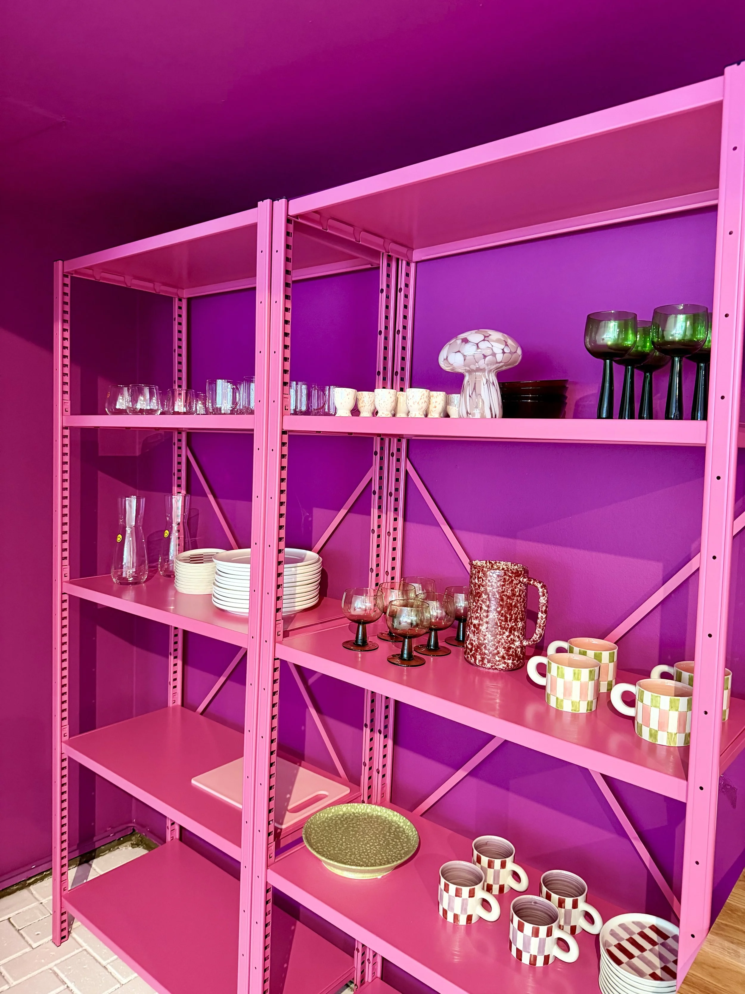 Pink metal shelf with glassware, cups, and decorative items against a pink wall.