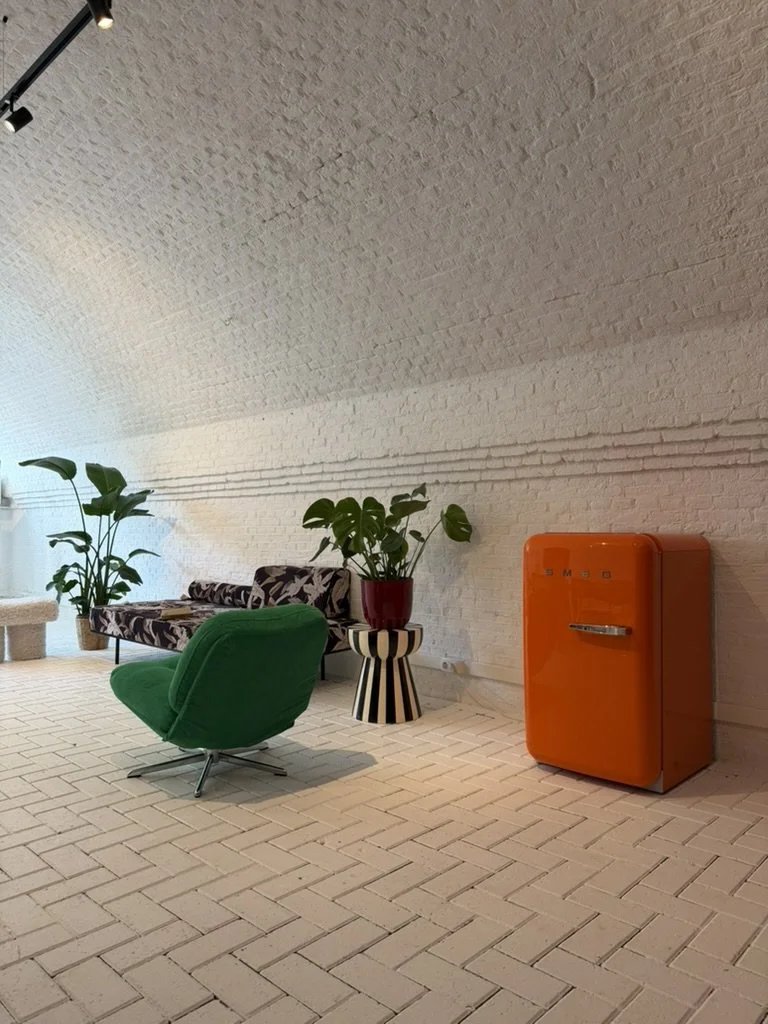 Modern interior with white brick walls and floor. Features a green armchair, a patterned sofa, potted plants, an orange vintage fridge, and a small striped side table.