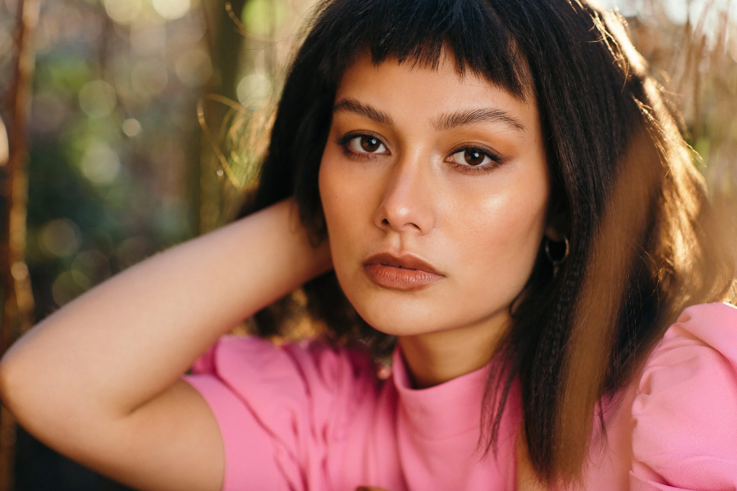 A young woman with medium brown skin and dark hair, partially covered by the hair with her arm raised, wearing a pink top, in a natural outdoor setting with sunlight filtering through the background.