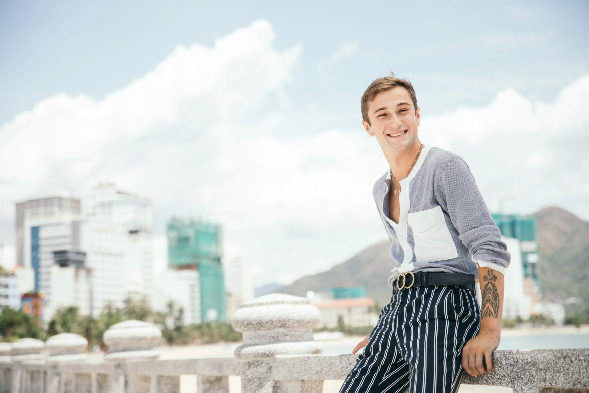 Young man outdoors smiling, wearing a striped shirt and striped pants, standing near a stone railing with a cityscape and mountains in the background.