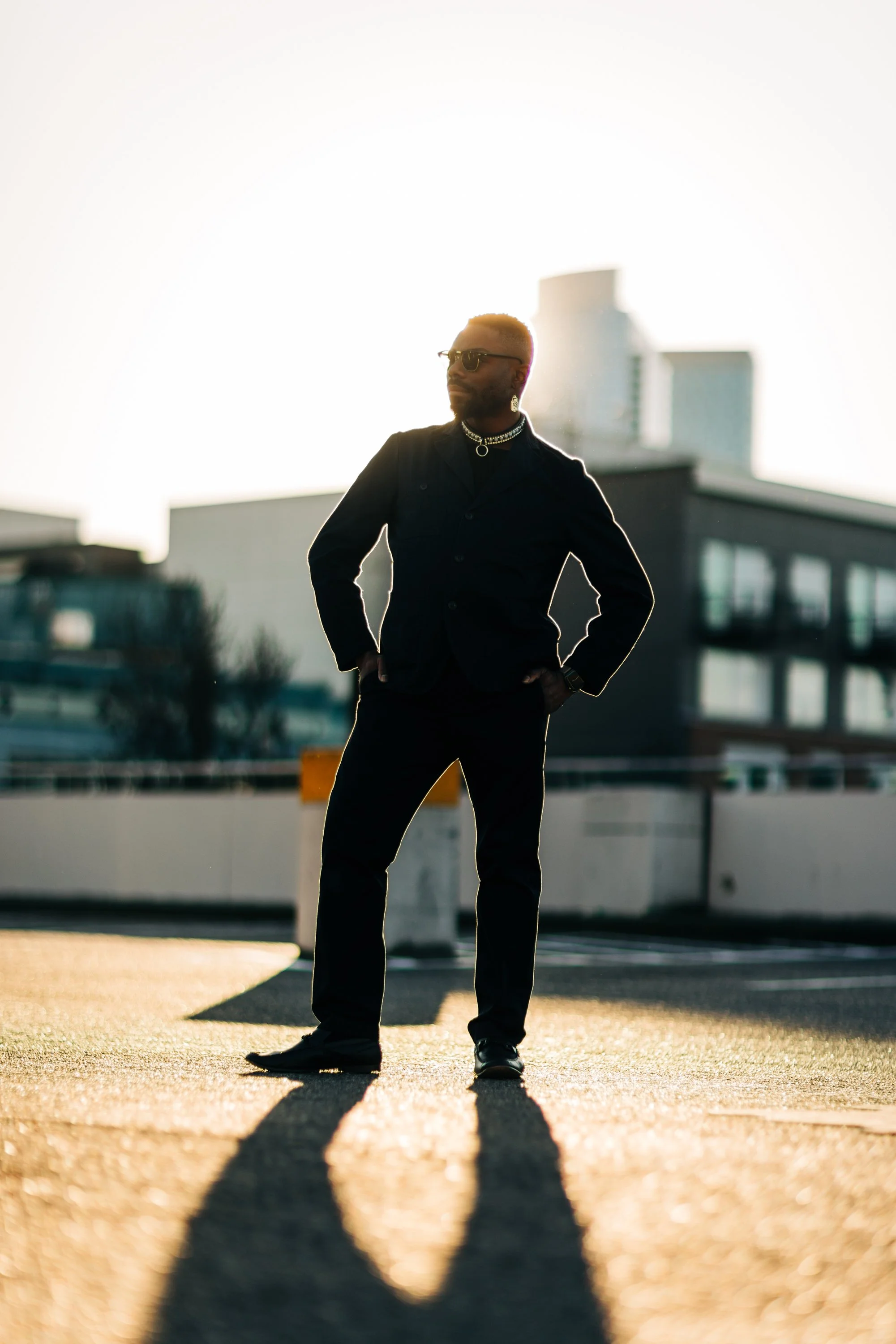 A man standing outdoors on a rooftop parking lot during sunset, wearing sunglasses, a black shirt, and jewelry, with a cityscape in the background.