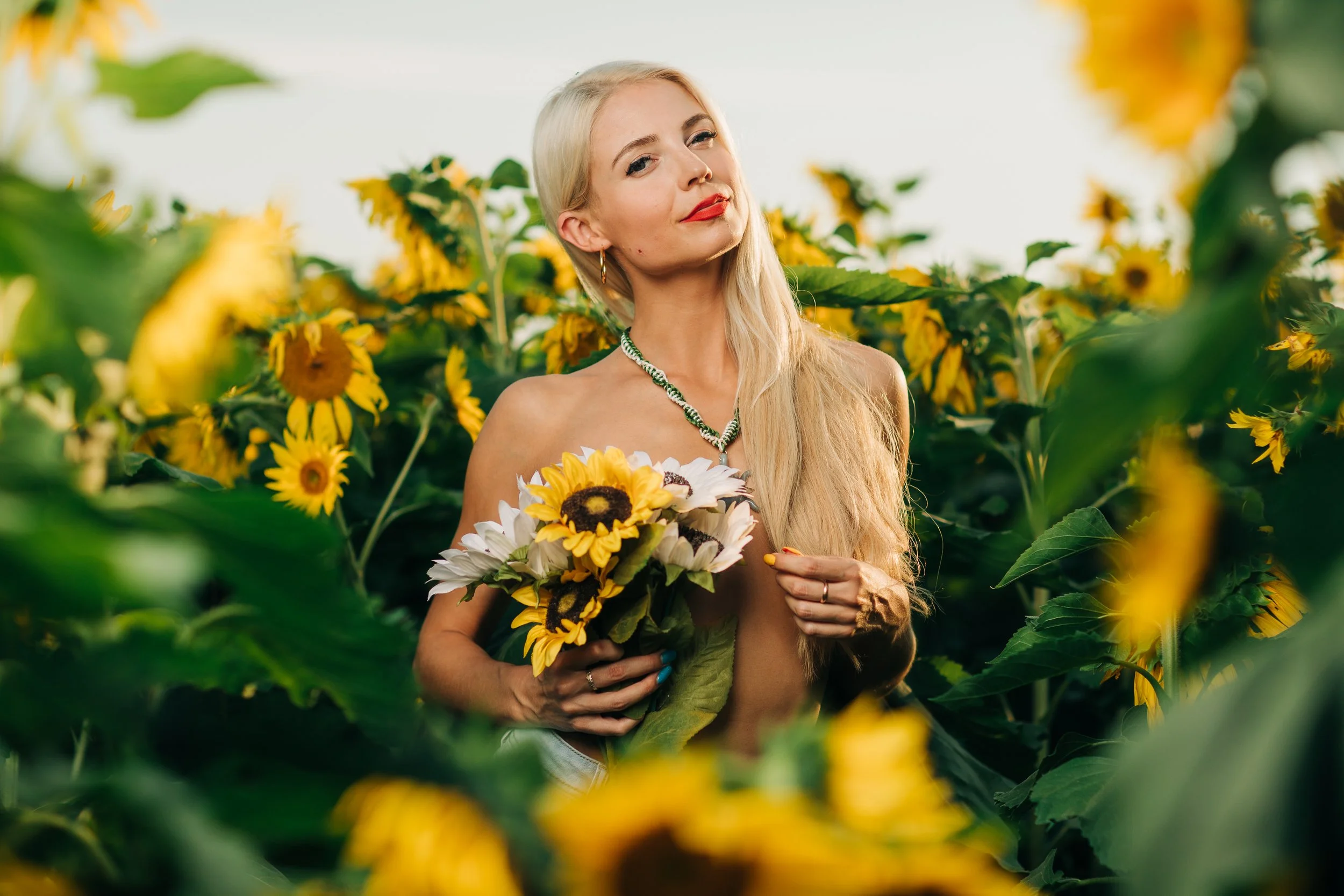 A woman with long blonde hair and red lipstick holding a bouquet of sunflowers and white flowers in a sunflower field.