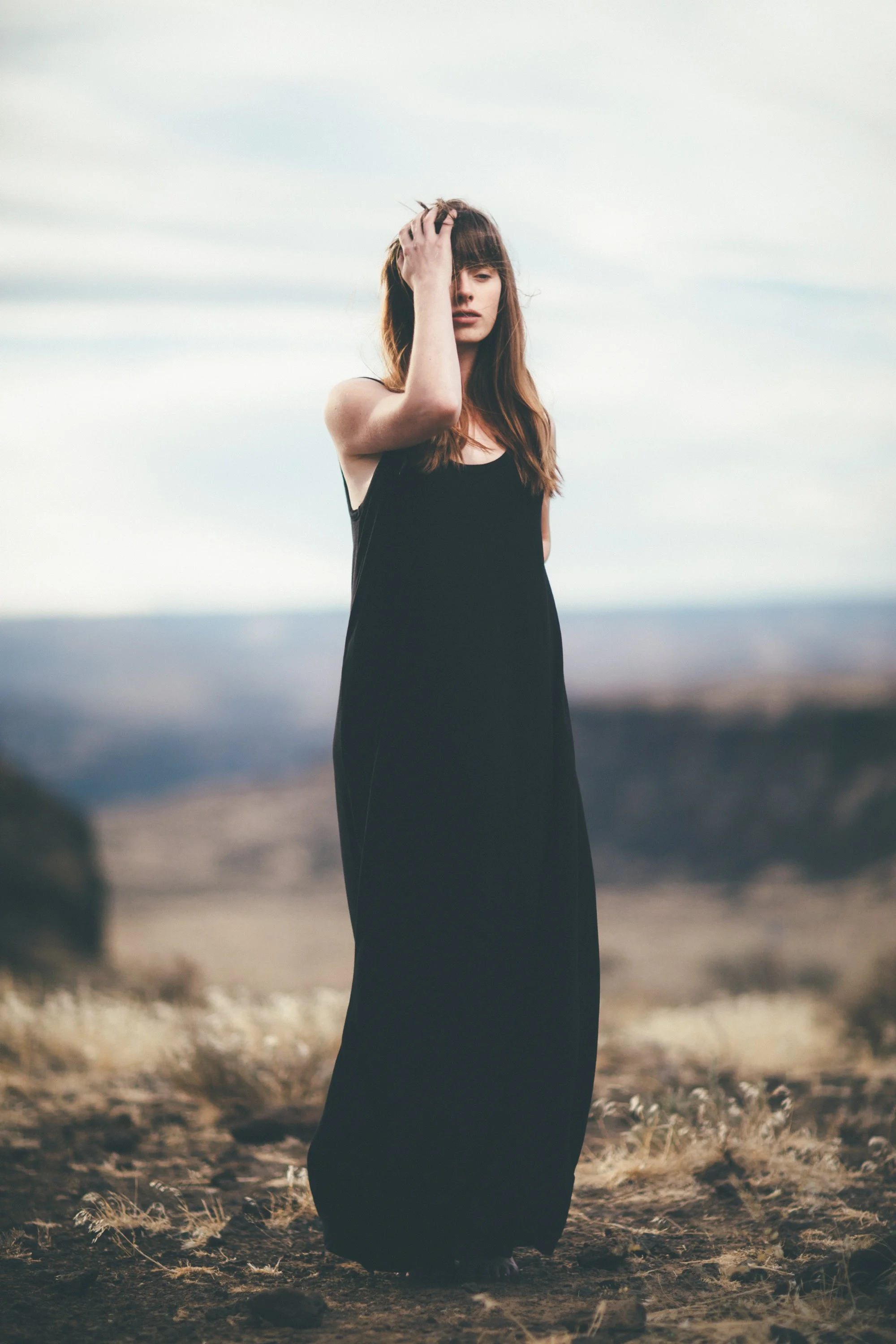 A woman standing outdoors on rocky ground with her left hand touching her forehead, wearing a sleeveless black dress, under a cloudy sky.