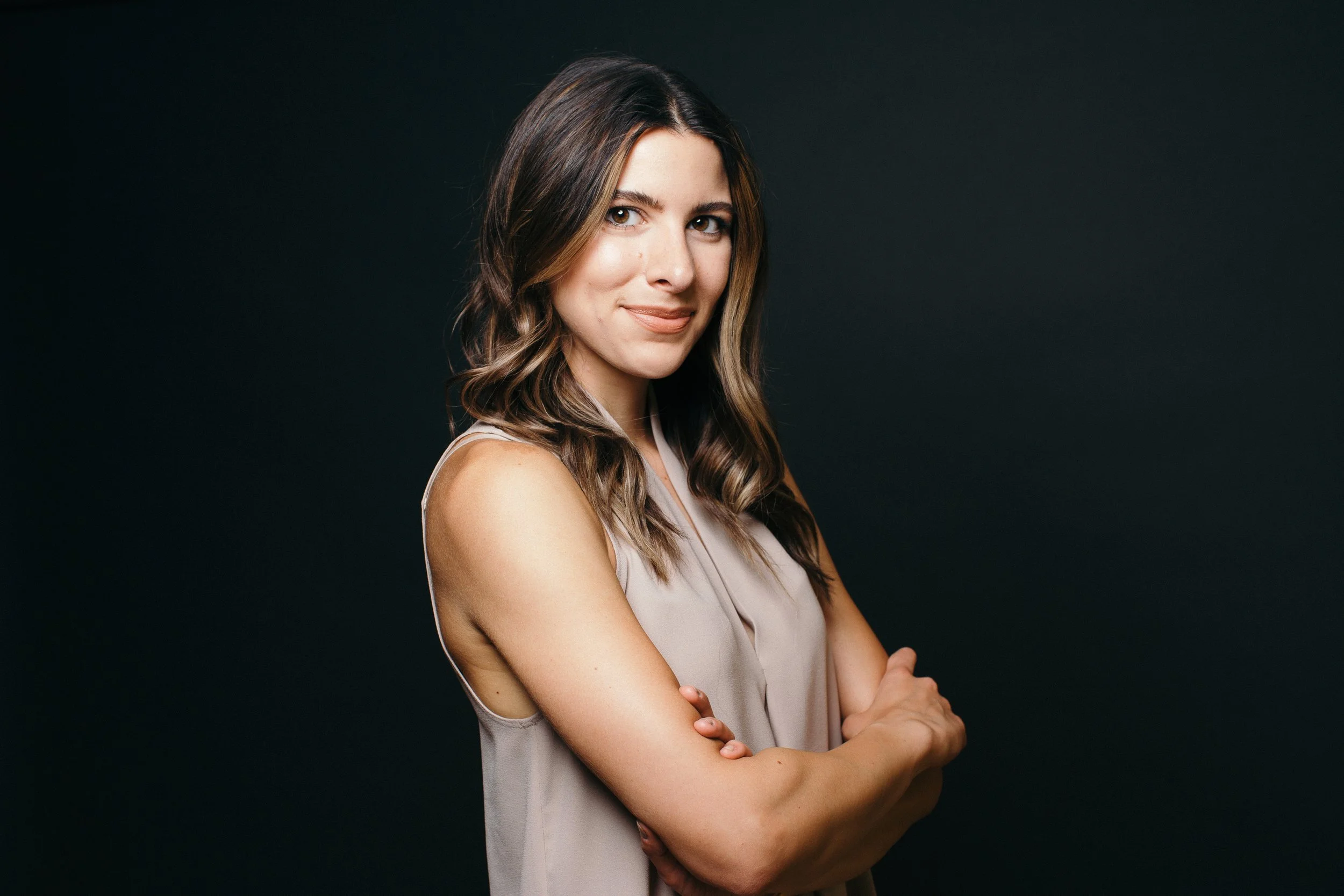 Portrait of a woman with brown hair, wearing a sleeveless beige top, standing with arms crossed against a black background.