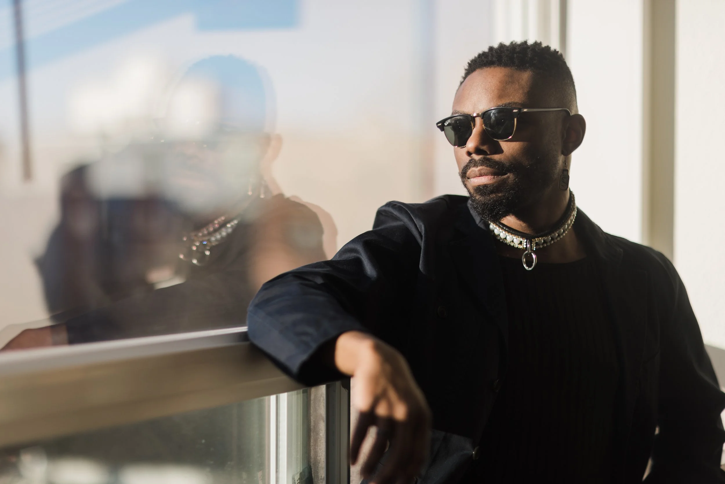 A man with short curly hair, beard, wearing sunglasses, a black outfit with jewelry, looking to the side, standing next to a reflective surface that shows his reflection.