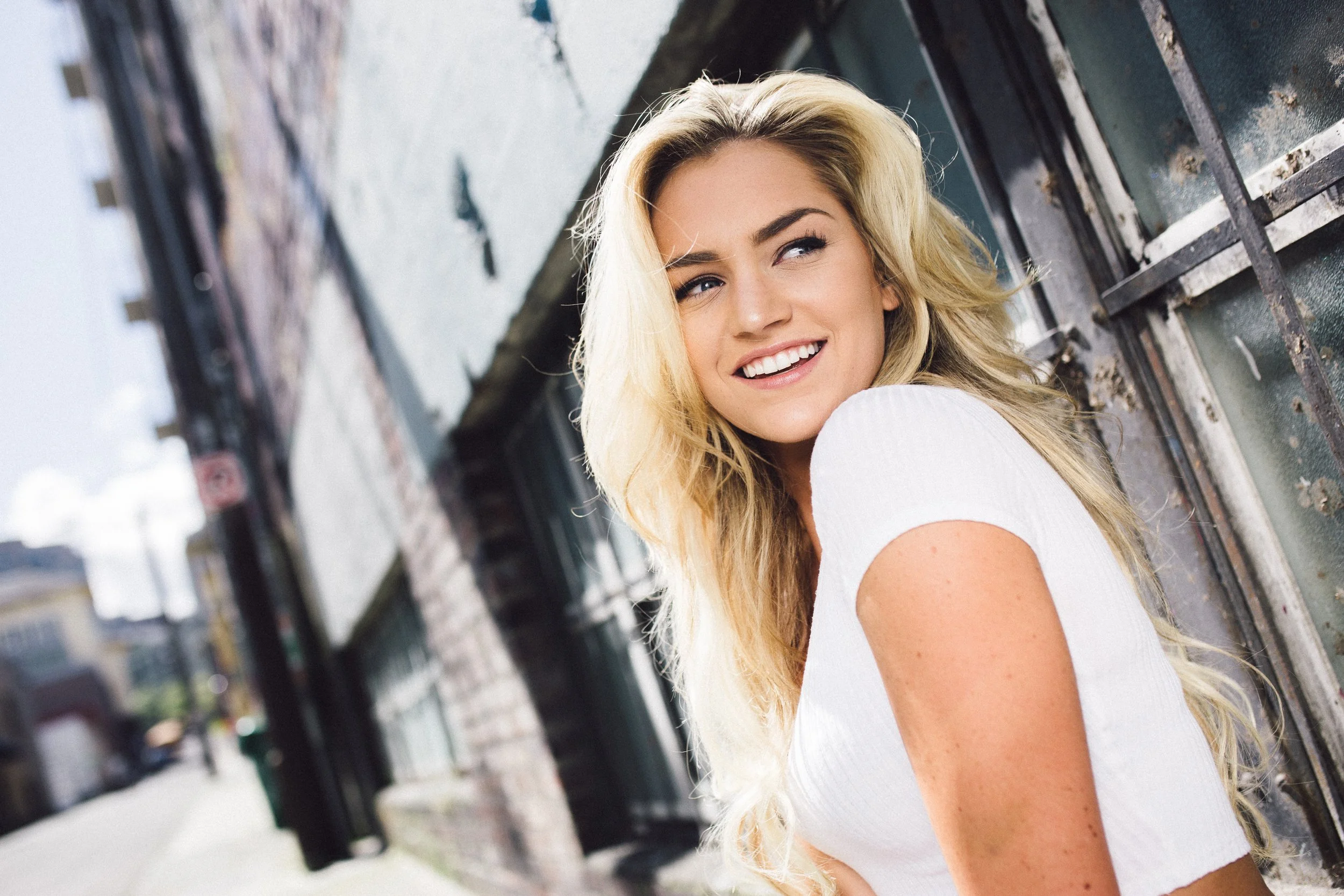 A young woman with long blonde hair and a white top, smiling and looking to the side, standing outdoors near a weathered metal and glass structure with a blurred cityscape in the background.