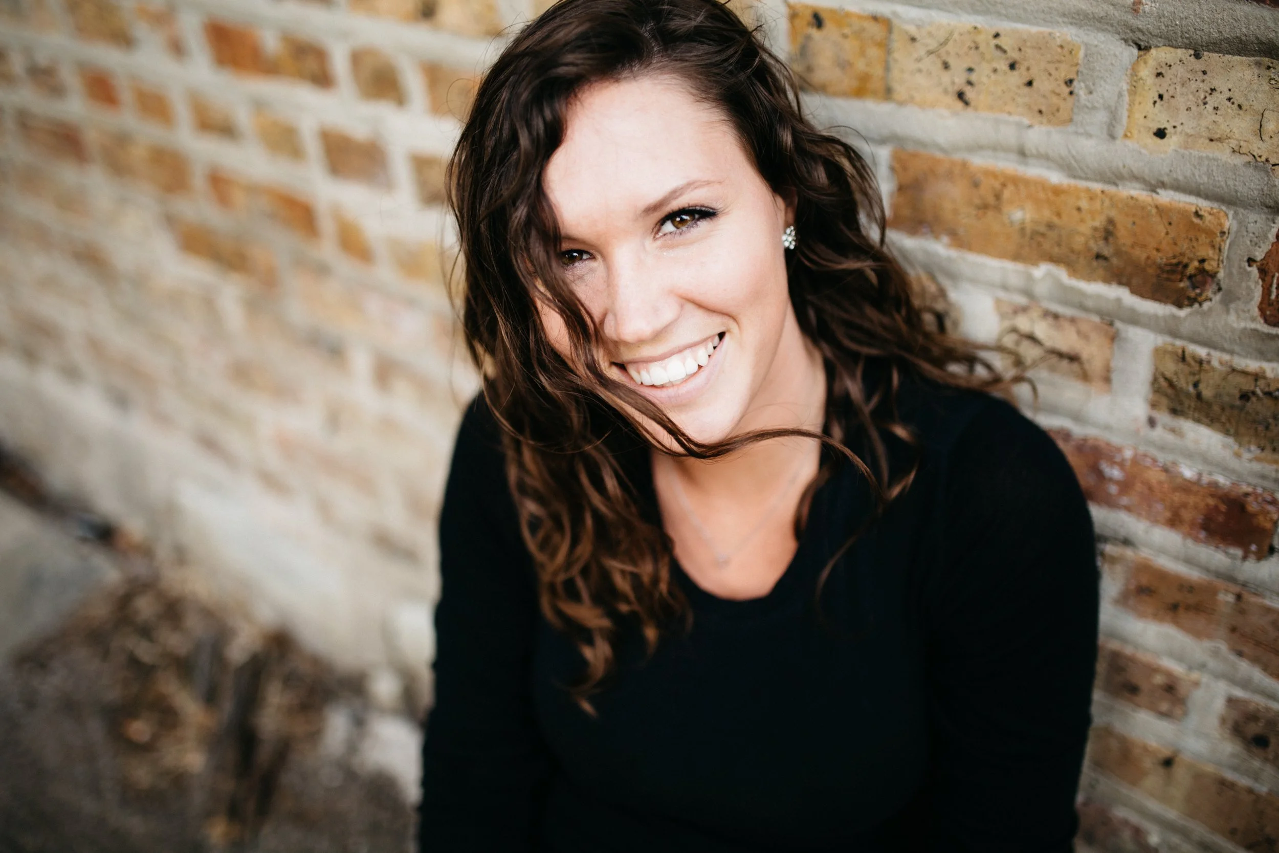 A young woman with curly brown hair, wearing a black top and floral earrings, smiling and leaning against a brick wall.