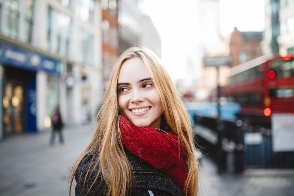 Young woman with long blonde hair smiling on city street near shops and red double-decker bus.