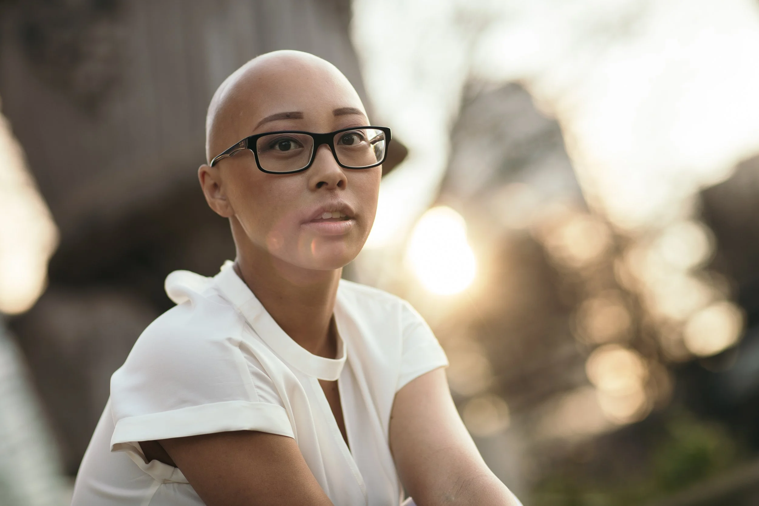 A woman with a shaved head wearing glasses and a white top outdoors with sunlight and blurred trees in the background.