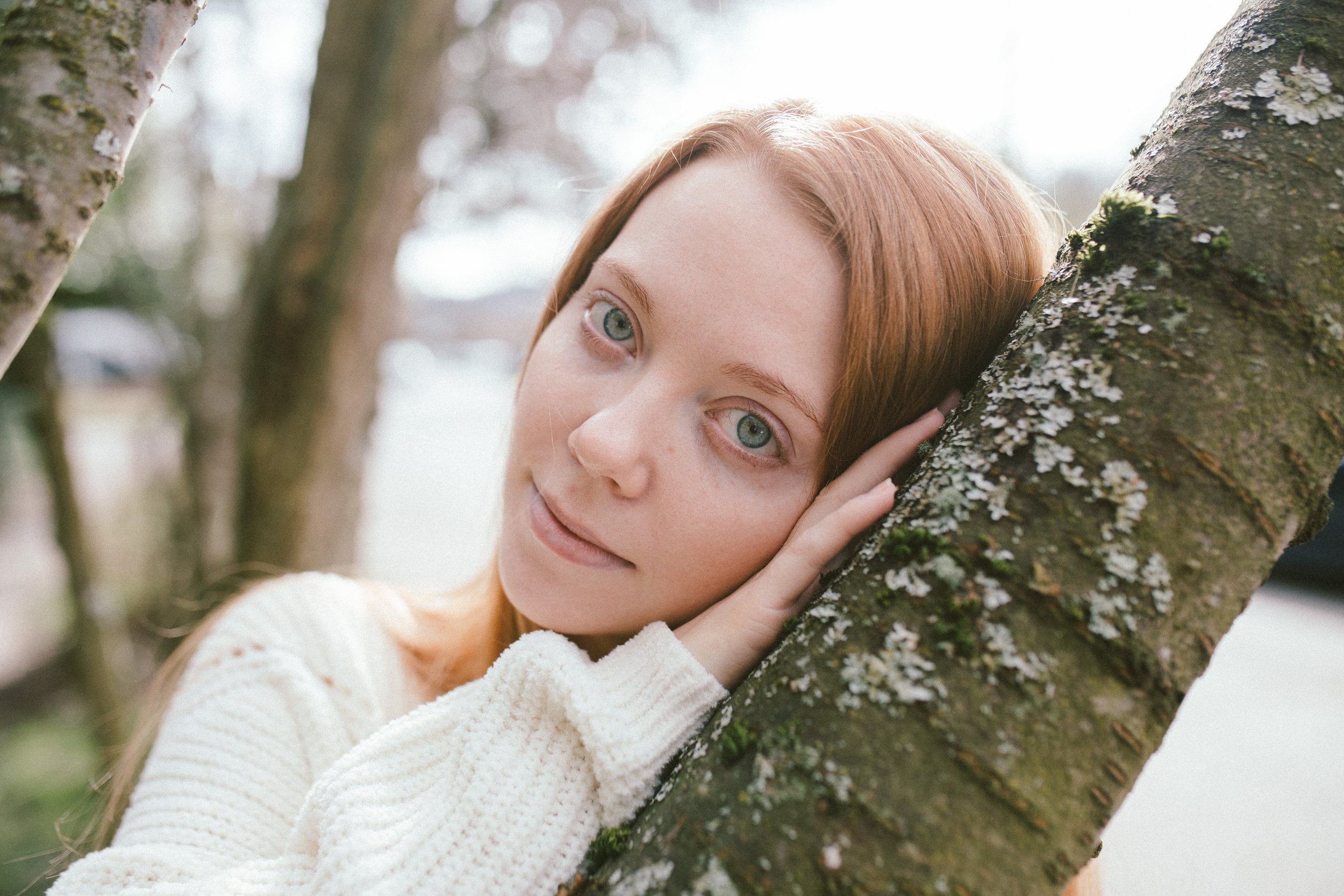 Young woman with red hair and blue eyes leaning against a moss-covered tree in an outdoor setting, wearing a white sweater.