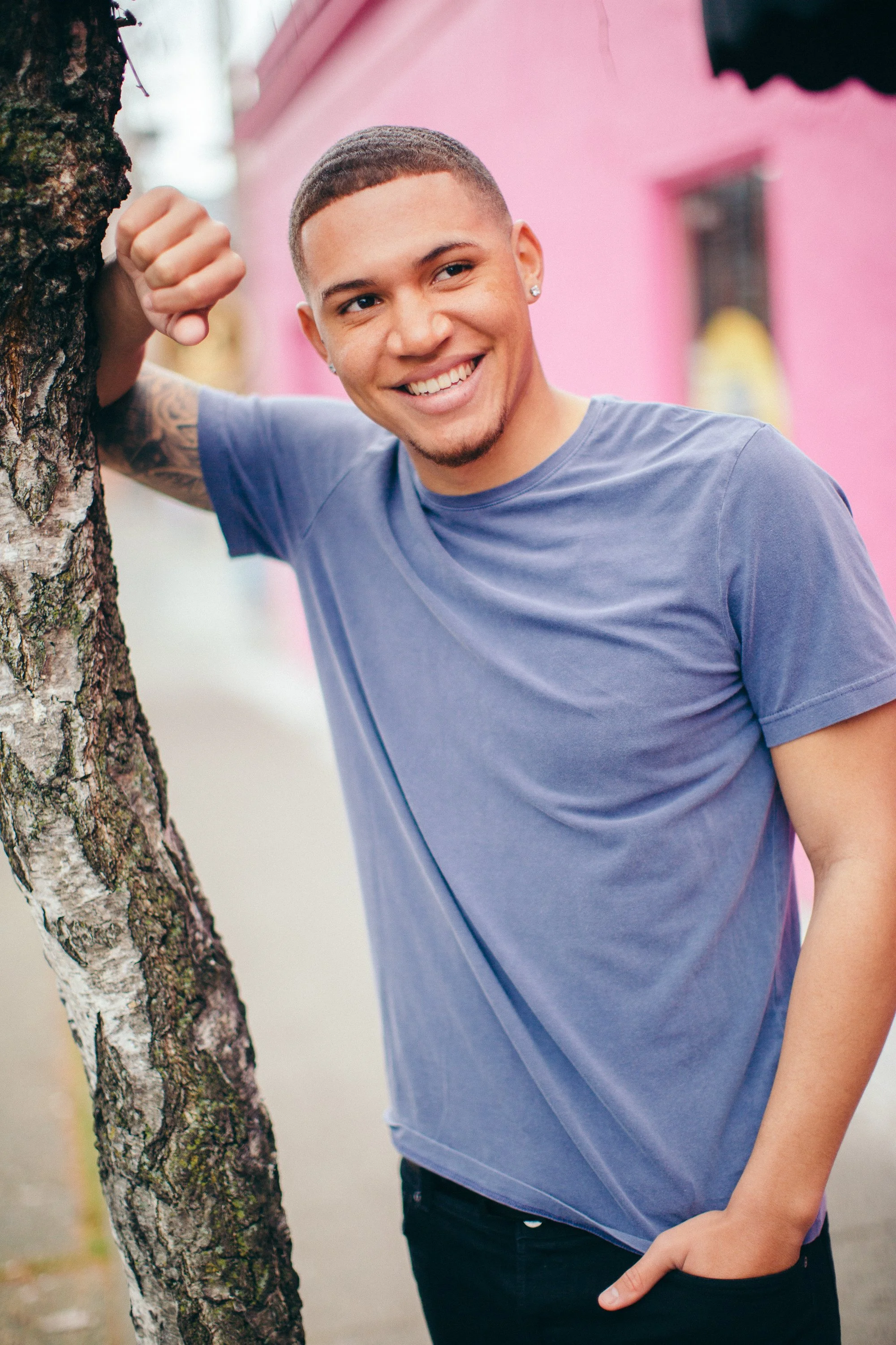 A young man with short hair and earrings smiling and leaning against a tree in an outdoor setting, wearing a blue t-shirt and black pants.