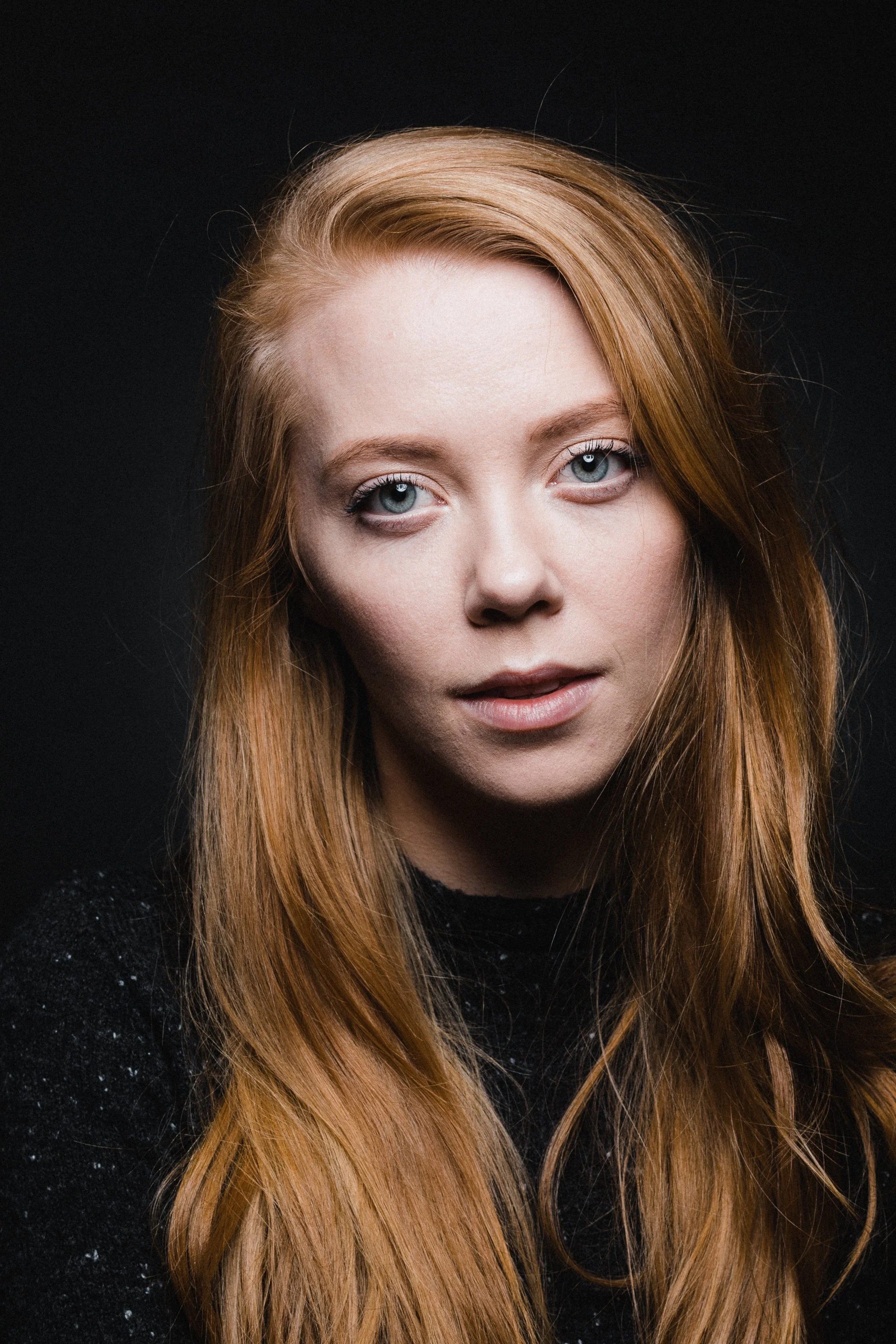 Close-up of a woman with red hair and light blue eyes, wearing a black sweater, against a dark background.