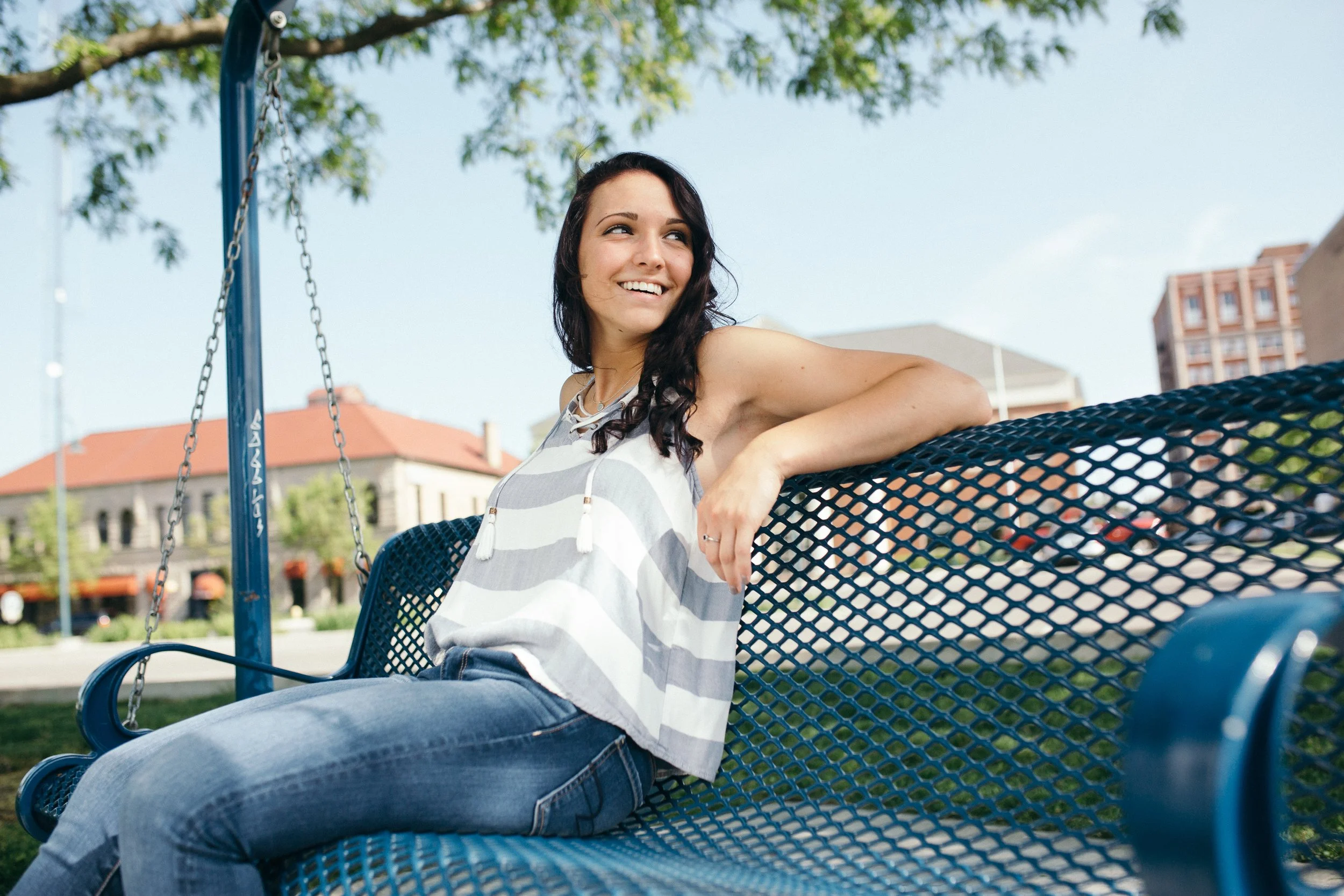 A woman with dark hair and a striped sleeveless top sitting on a blue playground bench, smiling and looking to the side, with a tree and buildings in the background on a sunny day.
