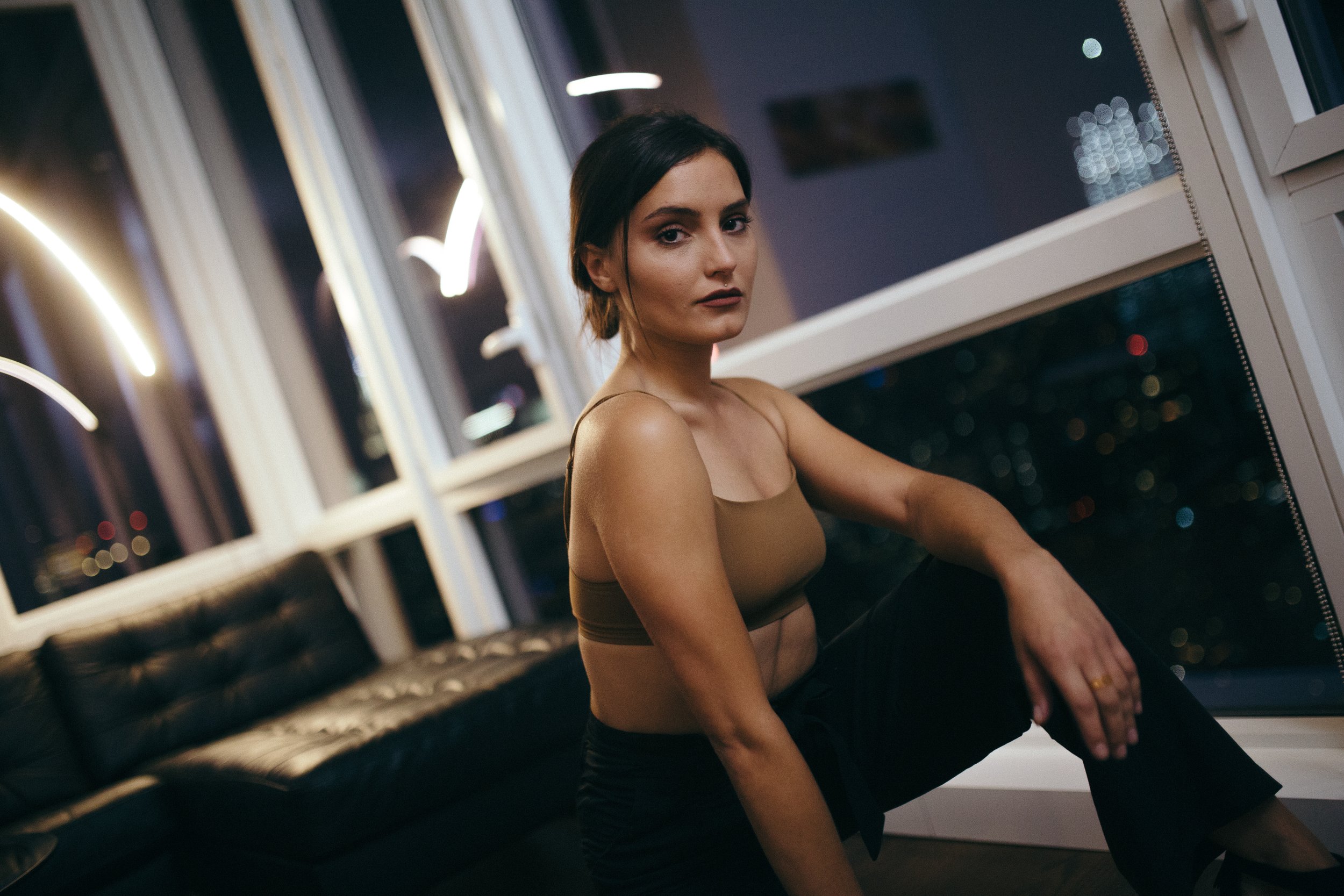 A young woman with dark hair and dressed in a brown top, sitting on a black leather couch in a modern room at night, with city lights visible through the window behind her.