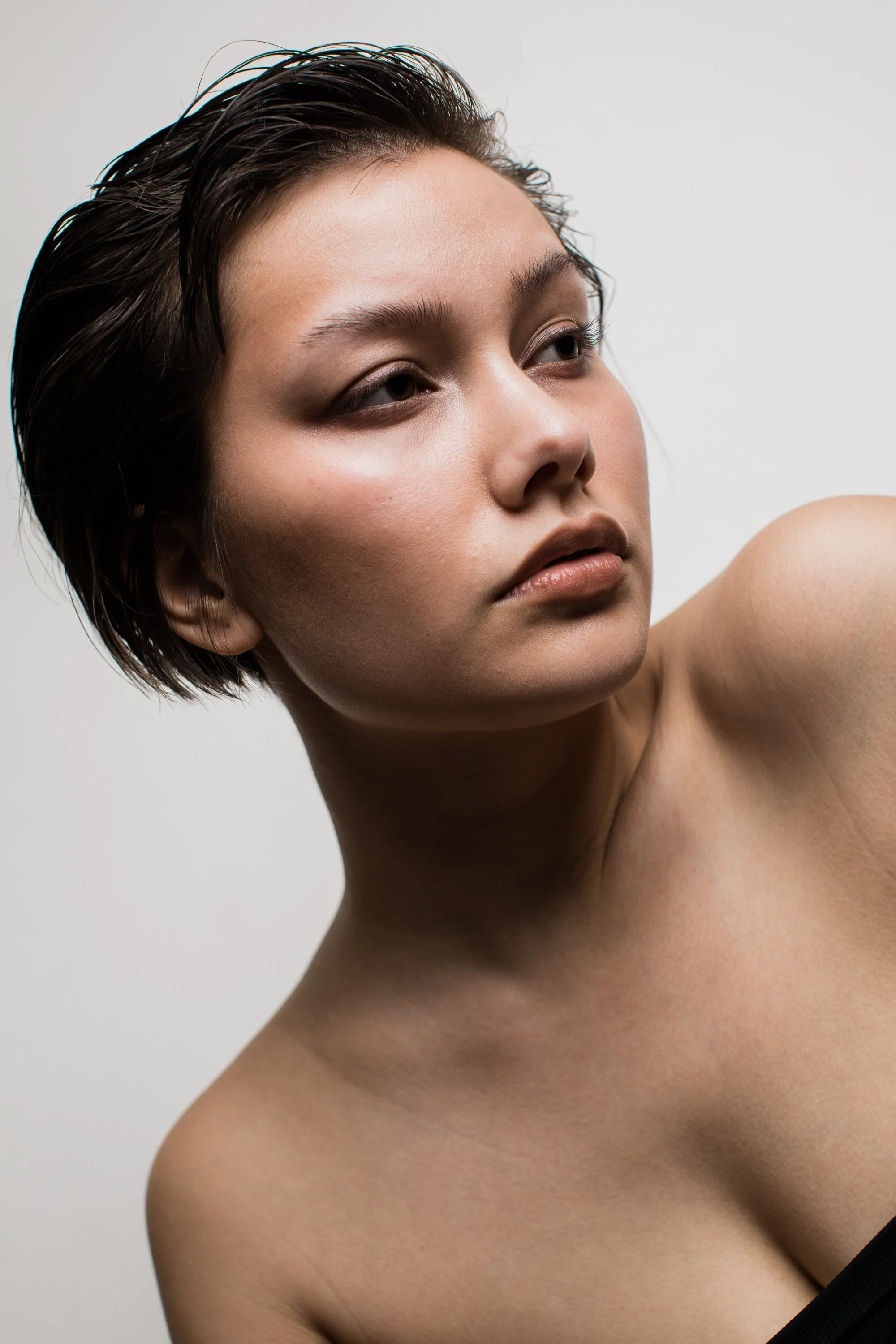 Close-up of a young woman with short wet hair, natural makeup, and clear skin, posing against a plain light background.