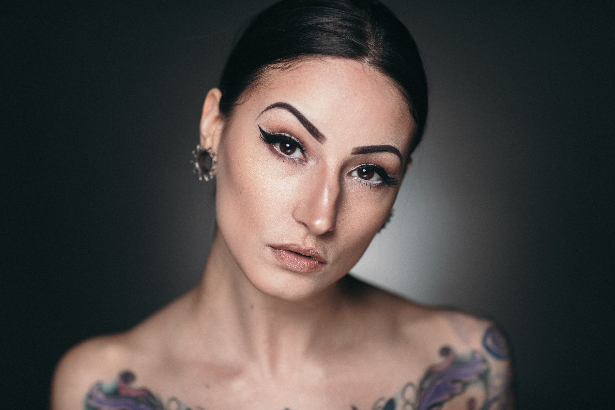 Close-up portrait of a woman with dark hair, bold makeup, and jewelry, against a dark background.