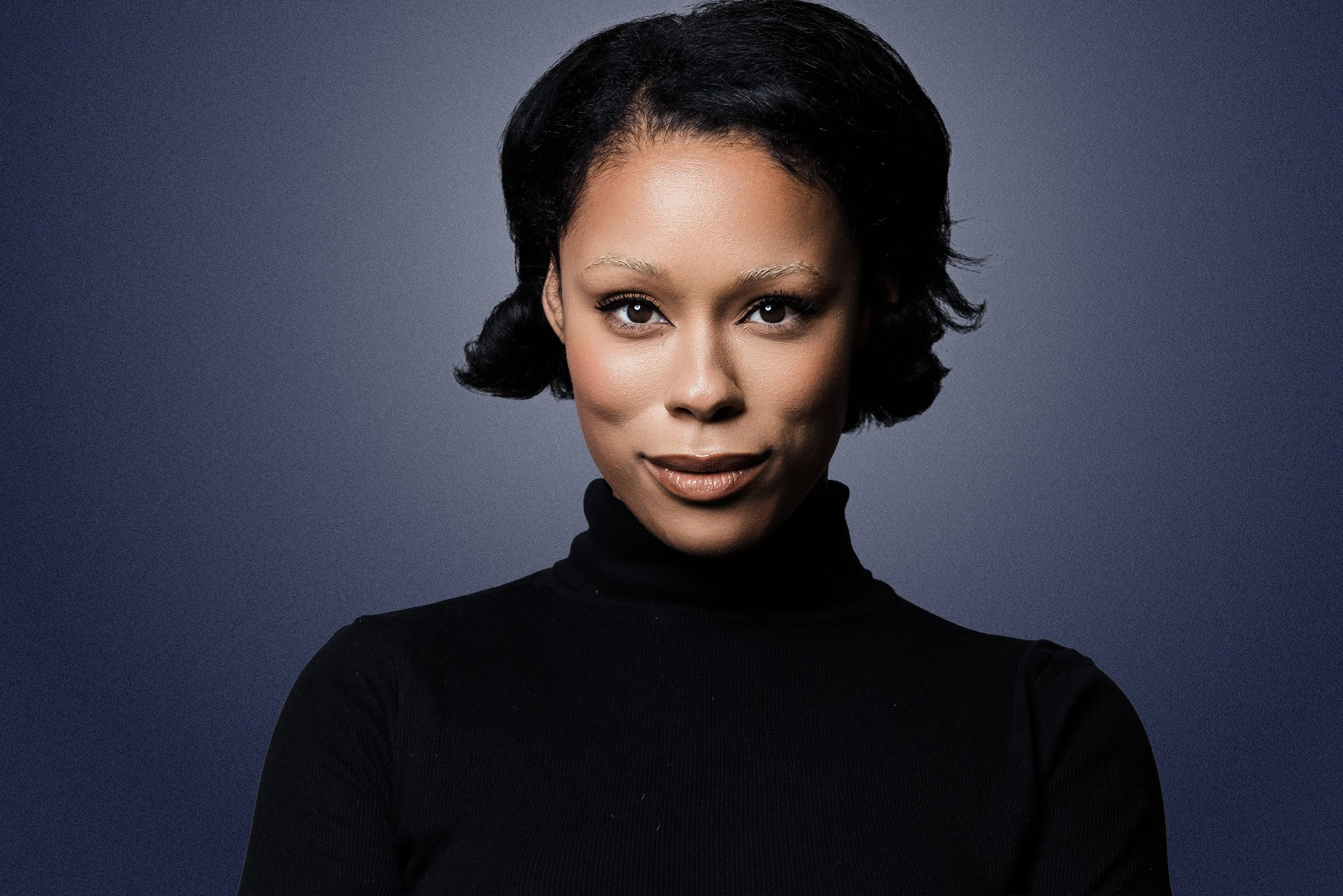 A woman wearing a black turtleneck, short black hair, and makeup against a plain gray background.