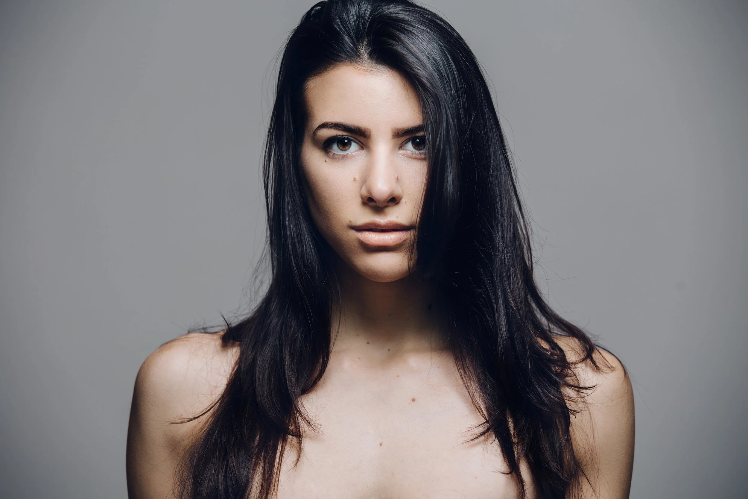 A woman with long dark hair, brown eyes, and a neutral facial expression poses against a plain gray background.