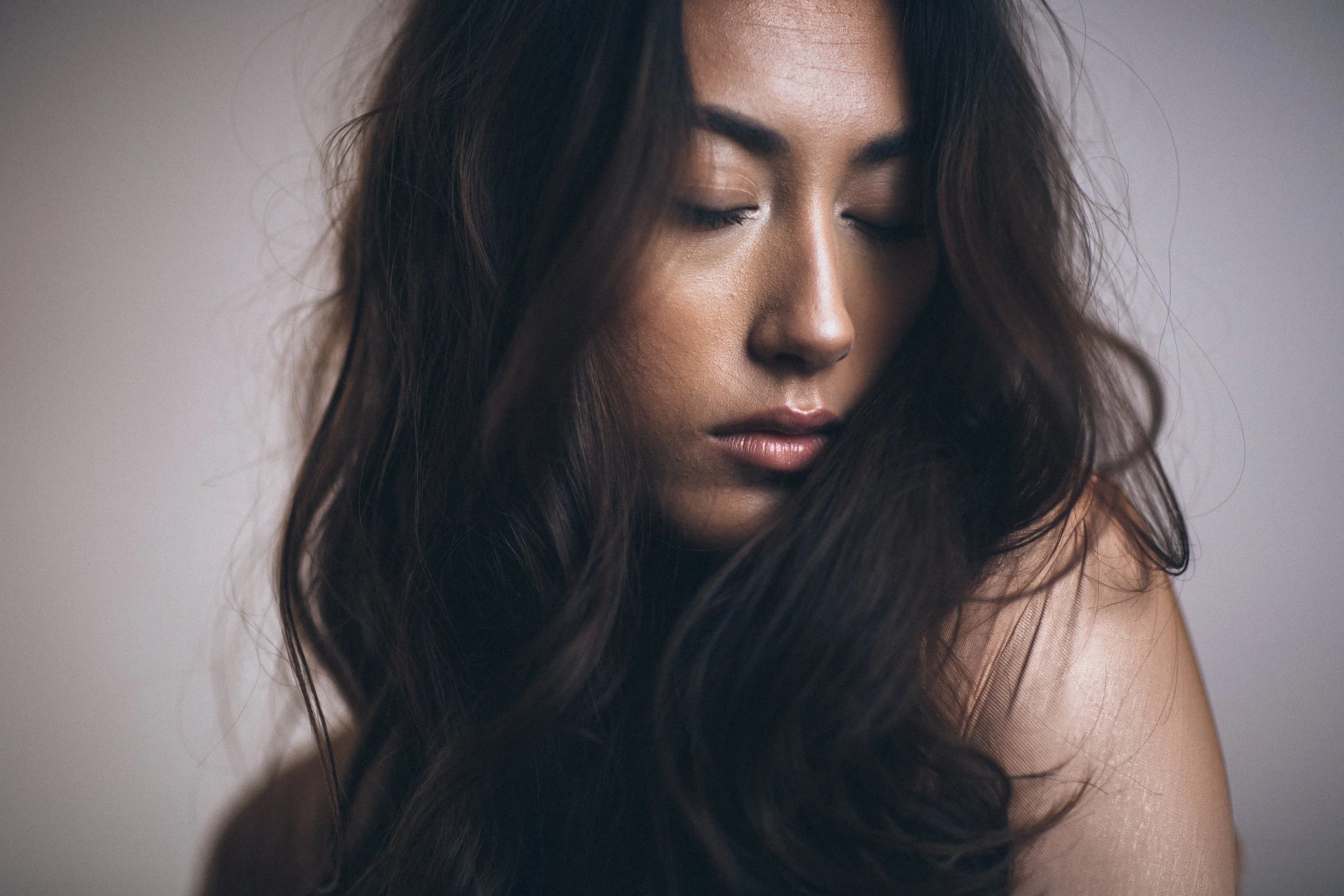 A close-up of a woman with long, dark hair and closed eyes, with soft lighting and a neutral background.