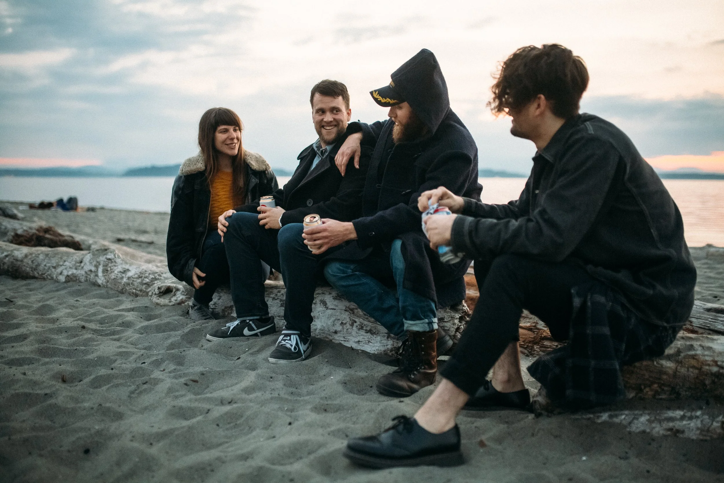 Four friends sitting on a log on the beach, chatting and smiling during sunset, with ocean and cloudy sky in the background.