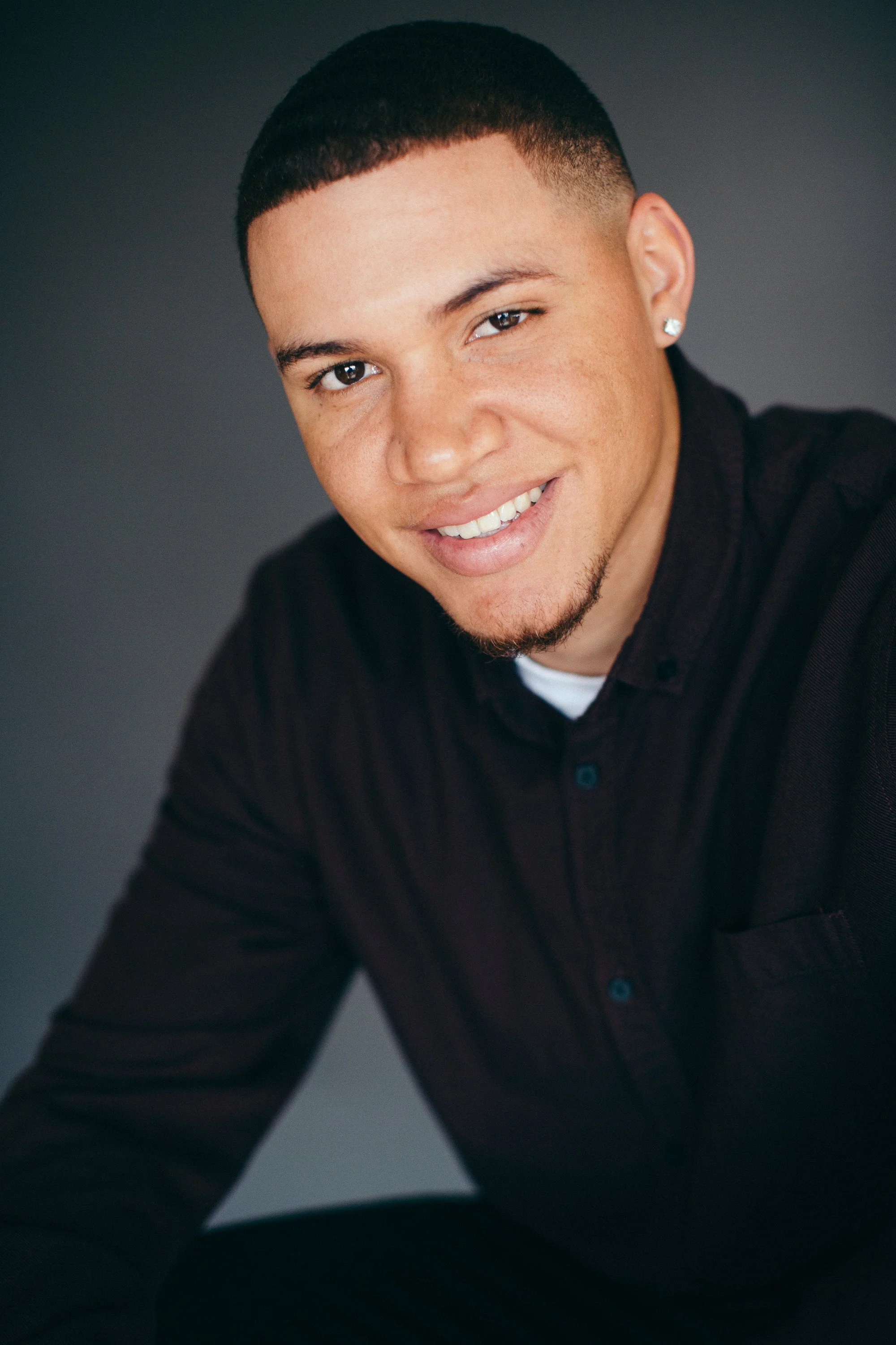 A young man with a short haircut, earrings, and a warm smile, wearing a black shirt, sitting against a dark background.
