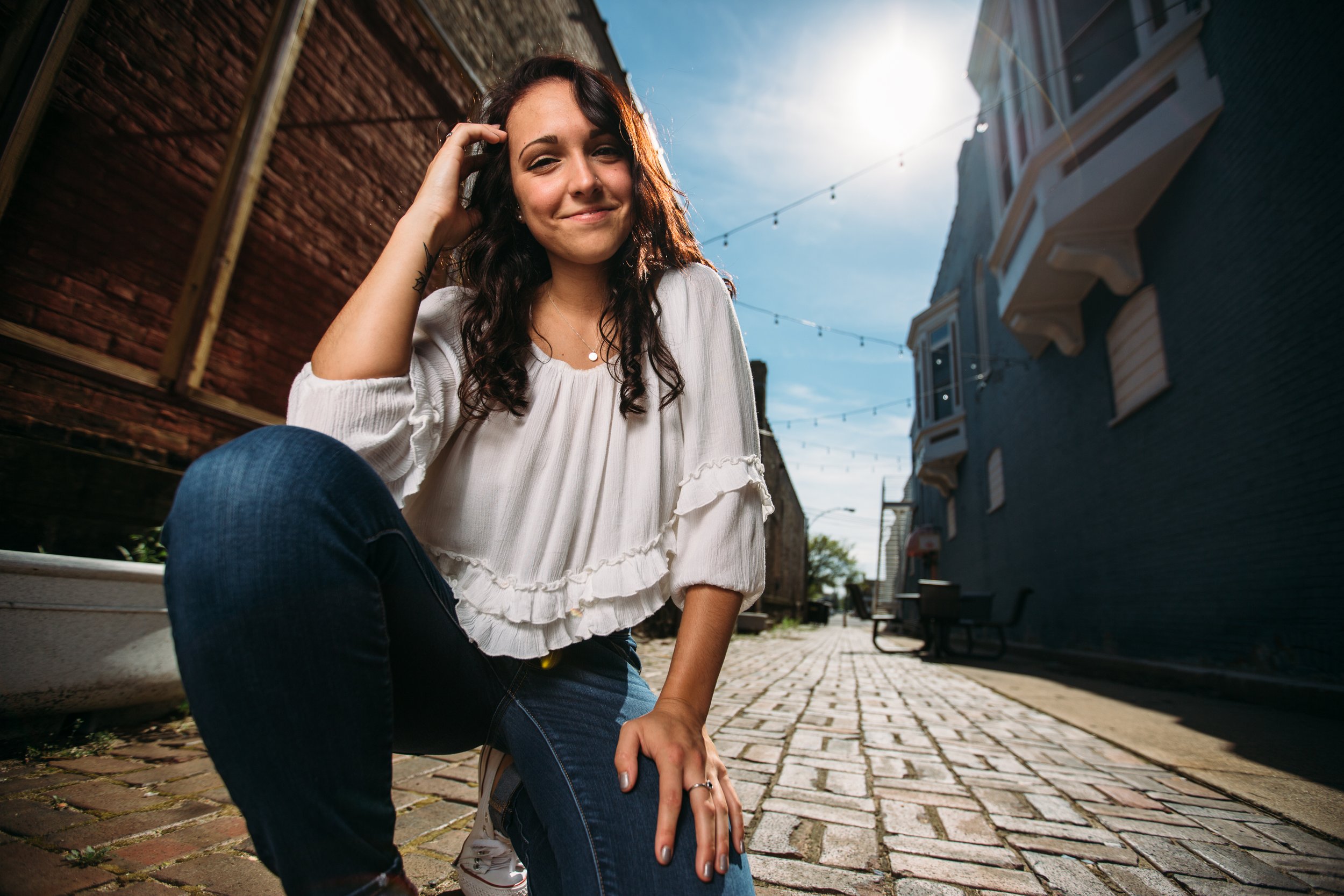 Young woman sitting on cobblestone street between brick and painted buildings, smiling at camera with sunlit sky in background.