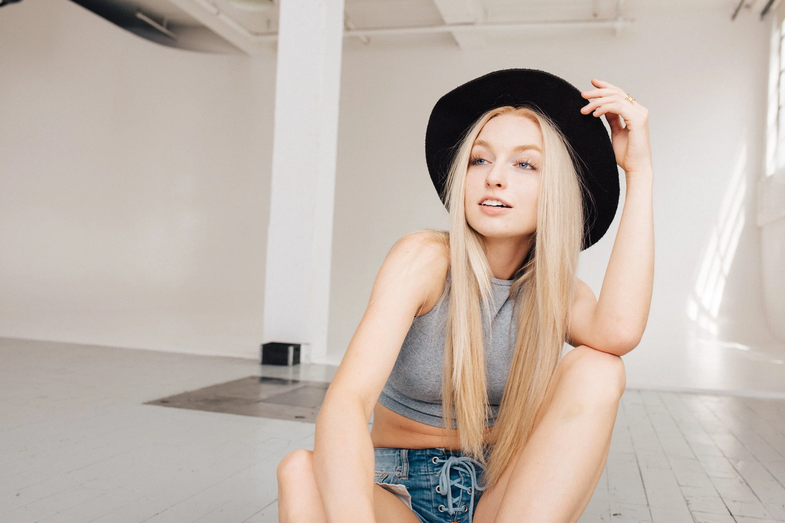 A young woman with long blonde hair wearing a black wide-brimmed hat, gray cropped top, and denim shorts sitting on the floor indoors, looking off to the side.